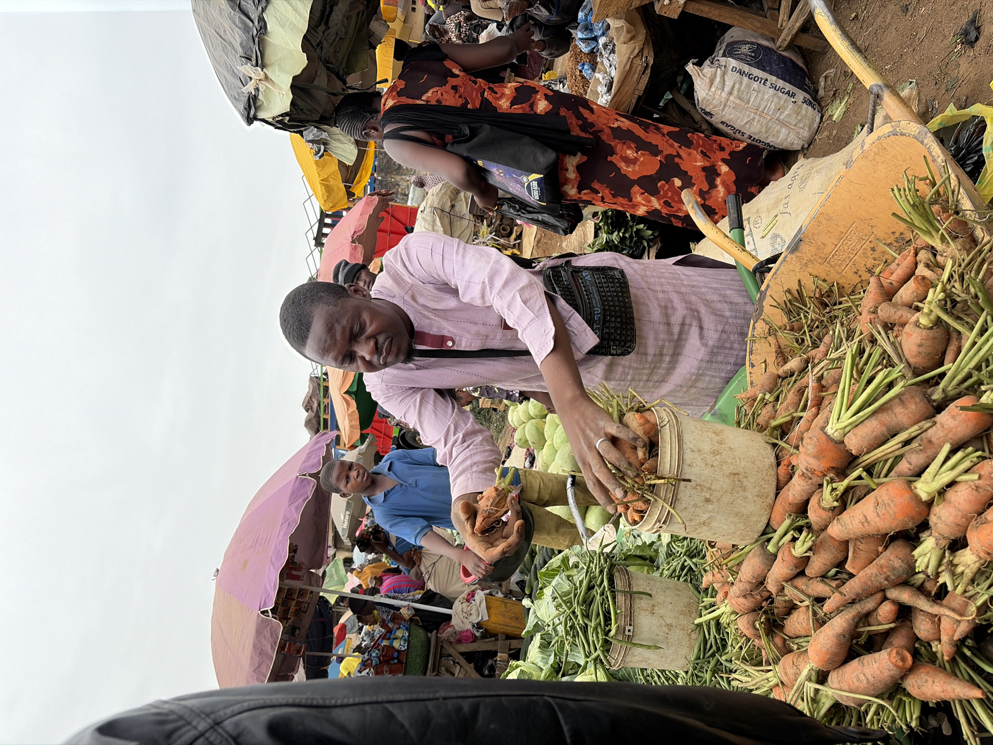 The image depicts an open-air market in Gura Top, Nigeria. A man wearing a light pink, long-sleeved shirt is selecting carrots from a white bucket, with a larger pile of carrots next to the bucket. Another man in a blue shirt stands nearby. A woman in a brown and orange floral dress and black bag is visible in the background. Other market stalls with umbrellas and goods are visible, including a bag of "Dangote Sugar." The scene is crowded with various items including cabbages and other produce. The sky is overcast.