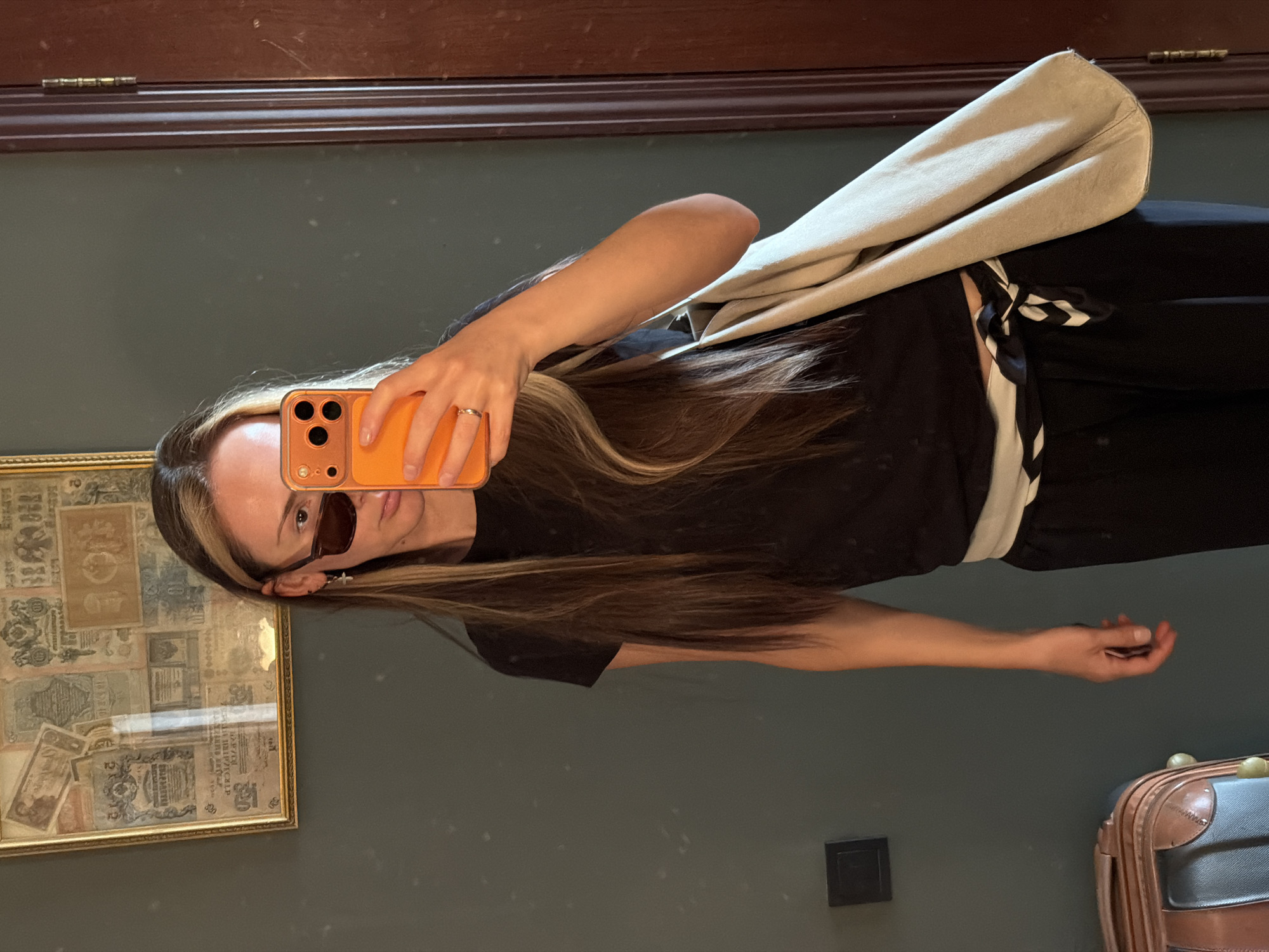 A woman takes a selfie in a mirror. She wears sunglasses and a ring. She is holding an orange phone in her right hand. She has long, dark hair with a blonde streak. She is wearing a black t-shirt and black pants with a white and black tie. She has a beige bag over her shoulder. Behind her is a green wall with a wooden frame at the top and a framed picture on the left. A power outlet is on the wall near the bottom. A suitcase is visible in the bottom right corner. The scene appears to be inside. There is no evidence to confirm the location is Khimki, Russia.