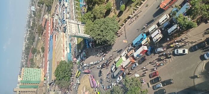 An elevated, wide-angle daytime photograph captures a segment of Landhies Road and the surrounding urban environment in Nairobi, Kenya, specifically overlooking the Machakos Bus Station (Muthurwa Bus Station) area and Nairobi City Stadium. The sky is clear to partly cloudy, casting distinct shadows.
The primary subject is a multi-lane asphalt roadway experiencing heavy vehicular congestion. Numerous public transport vehicles, including large buses and smaller matatus (minibuses) of diverse colors (blue, green, purple, yellow, red, white), are present, either stationary in queues or moving slowly. These are intermingled with private passenger cars (sedans, SUVs, vans) in various colors (black, white, red, silver). A large bus park or terminal area filled with additional parked public transport vehicles is visible in the middle ground, adjacent to the road.
Numerous pedestrians are observed on sidewalks, along road margins, and at crossing points. They appear to be walking, waiting, and congregating near bus loading zones.
In the background, a prominent multi-story building with a distinctive green-gridded facade is visible. Adjacent to the main road, the Nairobi City Stadium is identifiable by its white seating sections and a blue-roofed grandstand structure. Various other commercial and industrial buildings with mixed architectural styles and corrugated roofs are scattered throughout the mid-ground. A large, multi-panel billboard structure with a white framework stands on the right side of the main road. Several large, mature trees with dense green foliage are distributed along sidewalks and within traffic islands, providing greenery amidst the urban setting. The scene overall depicts a highly active and densely populated urban transportation hub.