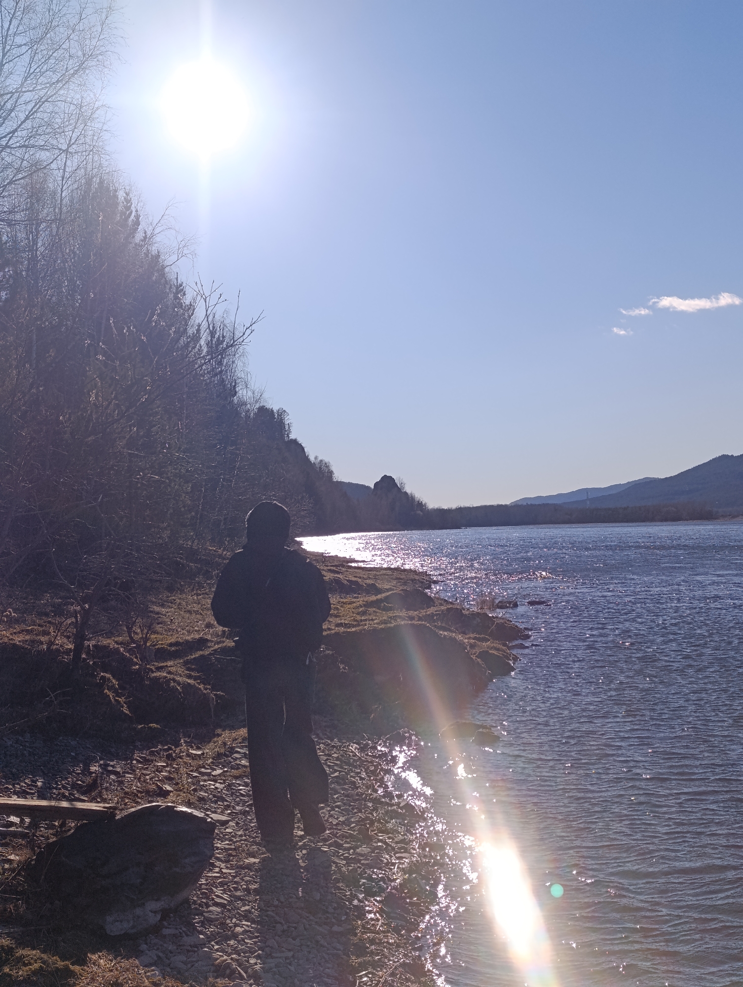 A person stands near the edge of the Yenisey River in Krasnoyarsk, Russia. The river reflects sunlight, causing glare on the water. The person is silhouetted and wearing dark clothing. Trees line the riverbank. In the distance, mountains are visible under a clear, blue sky with a single cloud. A prominent rock formation is on the horizon near the shoreline on the opposite bank of the river.
