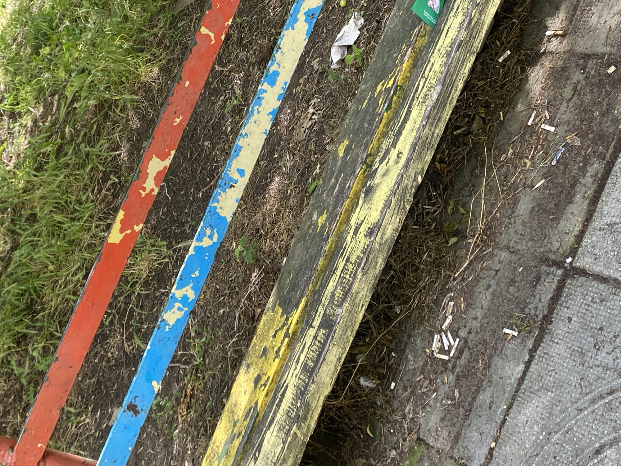 The image shows a close-up view of a weathered wooden park bench with peeling yellow paint. The bench is situated outdoors on what appears to be a paved area with dirt and dry leaves. Behind the bench, there are horizontal bars painted red and blue, also showing signs of wear. The ground around the bench is littered with numerous cigarette butts, some appearing to be recently discarded. A crumpled white tissue or paper is visible near the right edge of the bench. The overall scene suggests an outdoor public space in Varna, Bulgaria, that has not been recently cleaned.