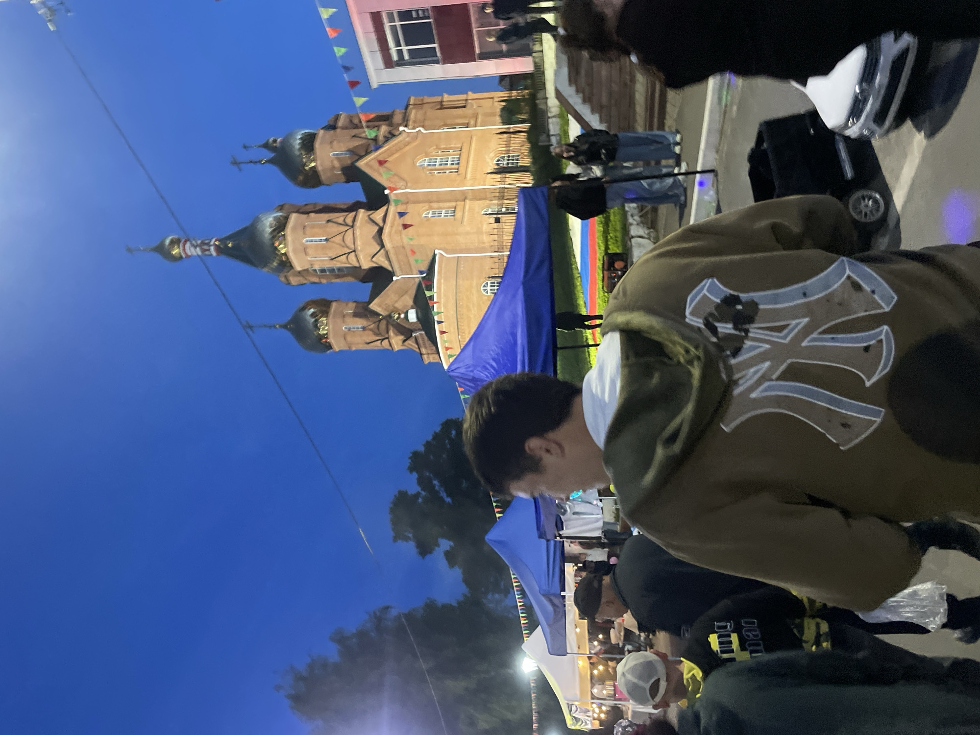 The image, taken in Красные Баки, Russia, shows an outdoor gathering at night. A large, orange church with multiple domes and a white building decorated with colorful flags are visible in the background. A blue tent is set up in the foreground, and several people are milling about. The focus is on a person wearing a brown jacket with a prominent "NY" logo on the back. Other individuals are partially visible, including one wearing a black shirt with yellow writing. Bright lights illuminate the scene, suggesting a festival or fair-like atmosphere.