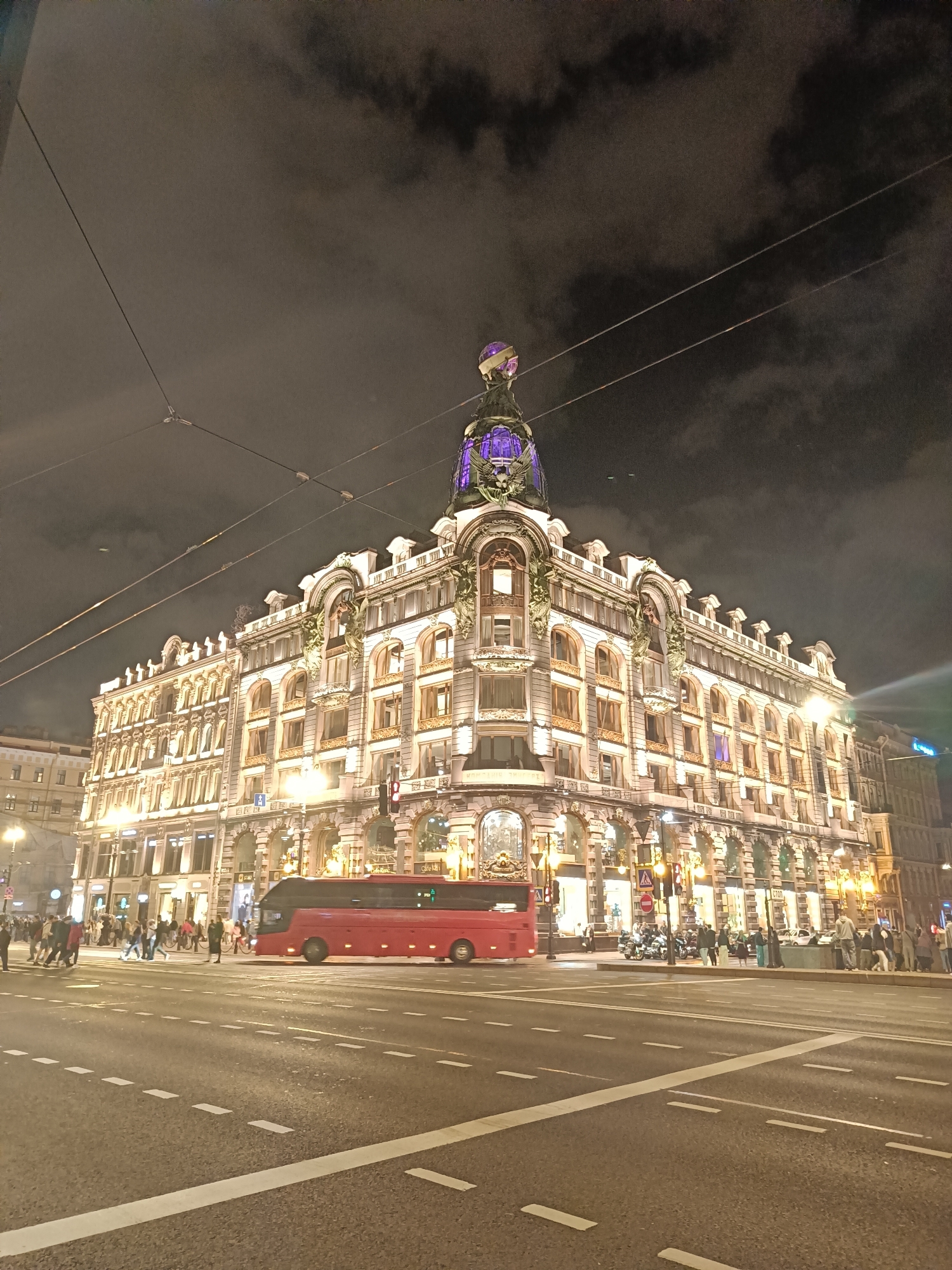 The image captures a night view of the Singer Building, also known as the Dom Knigi (House of Books), in Saint Petersburg, Russia. The architectural marvel is illuminated, highlighting its ornate facade and the distinctive glass dome topped with an eagle sculpture. A red tour bus is stopped at a crosswalk in the foreground, while pedestrians can be seen crossing the street and gathered on the sidewalks. The sky above is dark and cloudy, with power lines crisscrossing the frame, adding to the urban ambiance of the scene. The street is wet, suggesting recent rain.