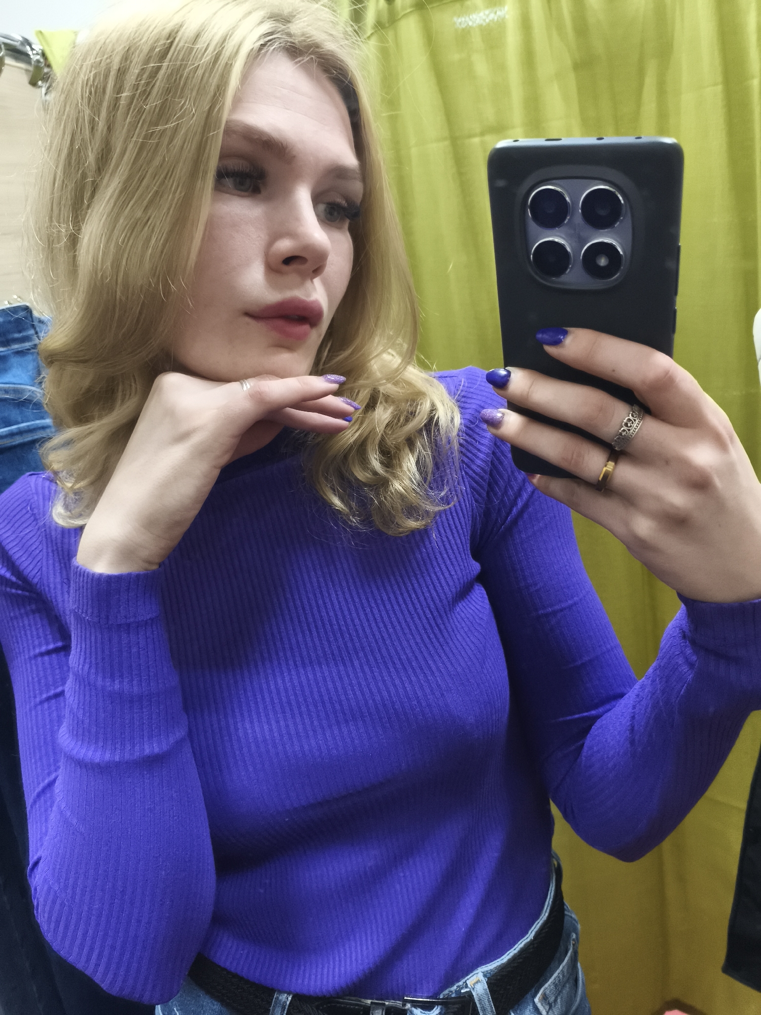 A young blonde woman is captured in a selfie taken in what appears to be a fitting room in Izhevsk, Russia. She has long, wavy hair and is wearing a vibrant purple ribbed long-sleeved top and blue jeans with a black belt. Her makeup includes defined eyeliner and plum-colored lipstick, and her nails are painted a deep purple, with glitter accents on her left pinky and ring fingers.
In her right hand, she holds a black smartphone with a distinctive module featuring four circular camera lenses arranged in a square. Her left hand is posed delicately under her chin, adorned with a silver ring on her pinky finger. Additional rings are visible on her right hand: a gold band on her ring finger and a silver patterned ring on her middle finger.
The background consists primarily of a chartreuse or olive green curtain, characteristic of a changing room. A glimpse of blue denim fabric, possibly another garment, can be seen on the far left. The woman's gaze is directed slightly away from the camera, giving her expression a pensive or posed quality, consistent with someone taking a self-portrait. There is no visible text in the image.