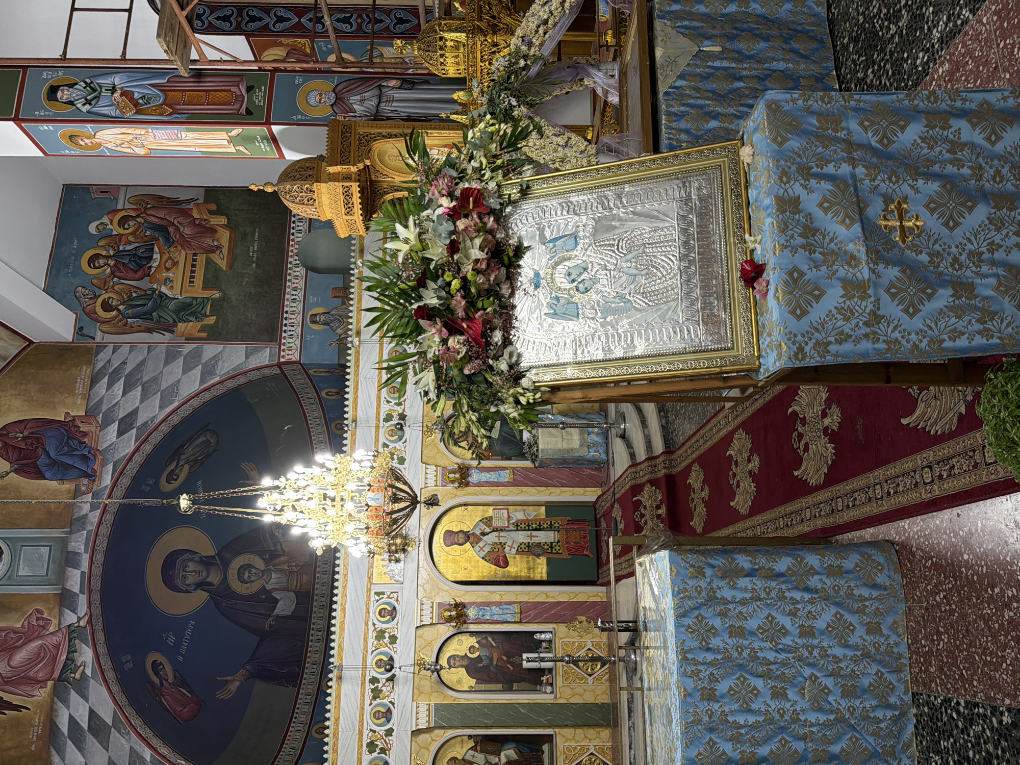 The image depicts the interior of an Eastern Orthodox church, likely located in the Municipal Unit of Alyzia, Greece. The scene is rich with religious iconography and liturgical elements. In the foreground, a large icon of the Virgin Mary and child, encased in a silver embossed frame, rests on a table draped with a blue cloth featuring a gold floral pattern. A vibrant floral arrangement crowns the icon. To the left, a red carpet with a gold eagle motif leads towards the altar area, flanked by tables covered in similar blue cloths. The background reveals a richly decorated sanctuary with numerous frescoes adorning the walls and ceiling, depicting biblical scenes and figures in the traditional Byzantine style. A grand chandelier hangs from the ceiling, illuminated. The overall impression is one of solemnity and religious devotion.