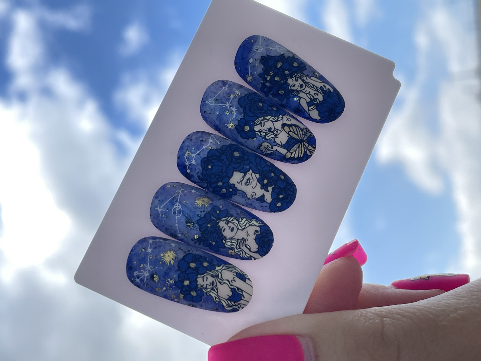 The image displays a white, rectangular nail swatch stick held against a bright blue sky with scattered white clouds, suggesting an outdoor setting or near a window in Izhevsk, Russia. The swatch stick holds five oval-shaped artificial nails, each featuring intricate hand-painted nail art.
The nail designs are predominantly dark blue with lighter blue and purple accents, creating a celestial or night sky theme. Each nail is adorned with white outline drawings of women, often depicted with elaborate, flowing blue floral hair that blends into the background. Gold and white speckles, resembling stars or stardust, are scattered across the nails. Visible constellation patterns and zodiac symbols include Cancer (♋) on the top nail and Libra (♎) on one of the middle nails. Specific designs also include a woman with a butterfly on her hand and another nail showing two women's faces intertwined.
A hand with bright, glossy pink manicured nails is visible in the bottom right, holding the swatch stick. The clear, bright sky indicates that the photo was taken during the daytime under good weather conditions. The overall impression is a showcase of detailed and artistic custom nail designs with a mythological and celestial theme.