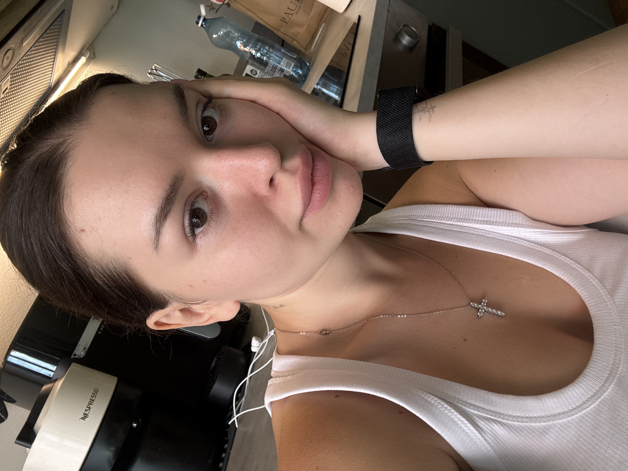 A young woman with brown hair pulled back leans her head on her right hand, looking at the camera. She is wearing a white ribbed tank top and a silver necklace with a cross pendant. A black watch is on her left wrist, and a faint tattoo is visible on her left forearm. In the background, a Nespresso coffee machine, a bottle of water, and a paper bag with "PAUL" printed on it are visible on a countertop. The environment appears to be a kitchen. No specific location in Prague, Czechia, can be determined from the image.