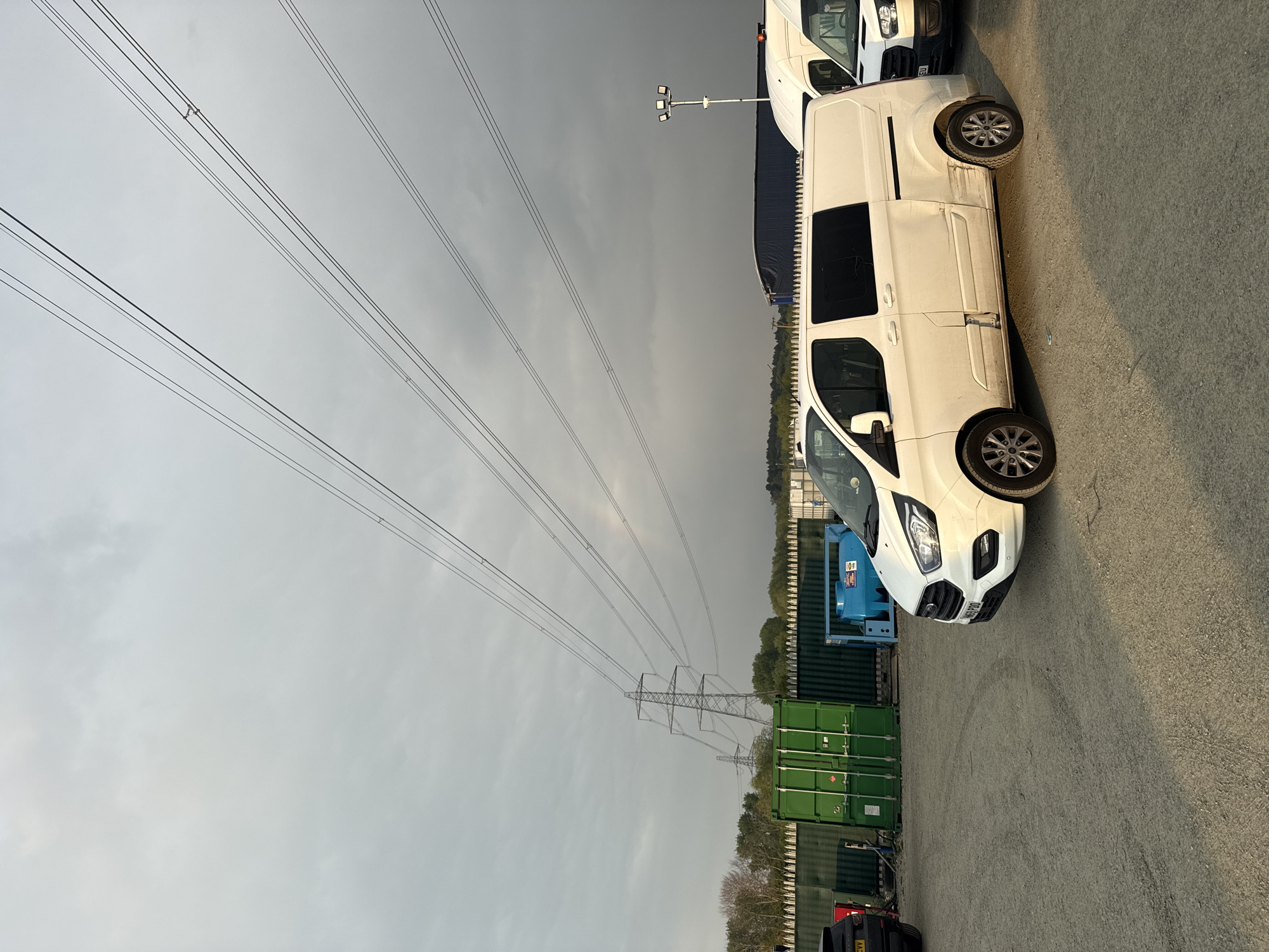 The image, oriented horizontally, captures an outdoor industrial or commercial yard under an overcast sky in Mid Suffolk, United Kingdom. The foreground shows an asphalt or gravel surface. Two white commercial vans, possibly Ford Transit Customs, are prominently parked. The closest van has the visible license plate "NG67 RDO". Another white van behind it features an "EU" sticker on its front grille. Further back, stacked green shipping containers form a boundary, behind which a line of trees is visible. In the mid-ground, a tall metallic lattice-work electricity pylon stands against the sky, with multiple power lines stretching diagonally across the frame. A second pylon is faintly visible in the far distance. A lamp post equipped with floodlights is also present near the vans. The sky is uniformly grey and cloudy, indicating overcast weather. No specific activity or individuals are depicted. A small black car with the license plate "MA22 CVW" is partially visible in the bottom left corner. The overall scene suggests a quiet, possibly late afternoon or early evening setting, given the soft, diffused light.