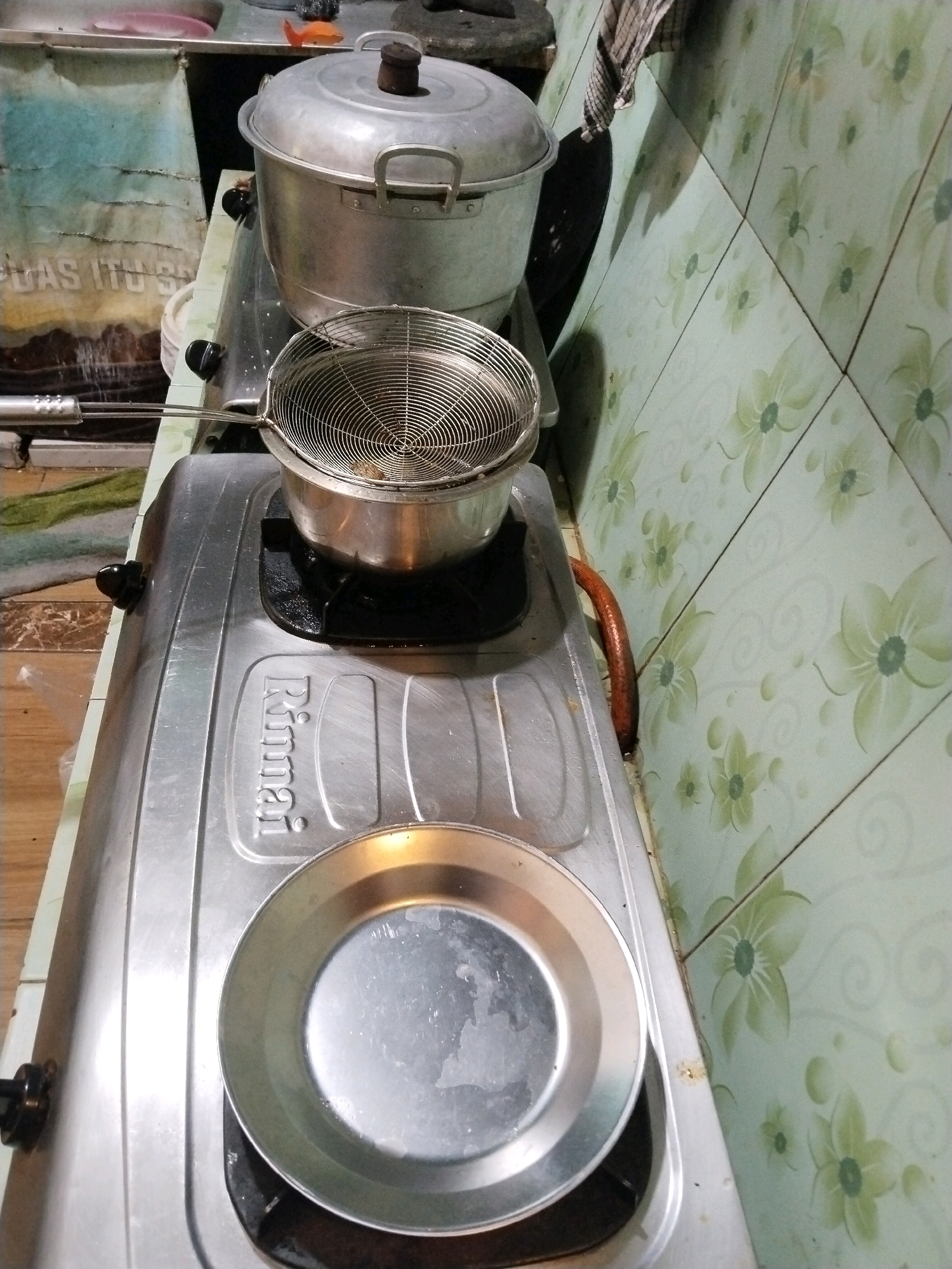 A slightly high-angle medium shot shows a stovetop in a kitchen in Prigi, Indonesia. The stovetop, made of aluminum, has two burners. The left burner is occupied by a large, aluminum pot with a lid. A wire strainer basket sits atop a smaller pot on the right burner. A small, metallic plate rests on the front of the stovetop. In the background, a tiled wall with a floral pattern is visible. The overall scene suggests a domestic cooking environment.