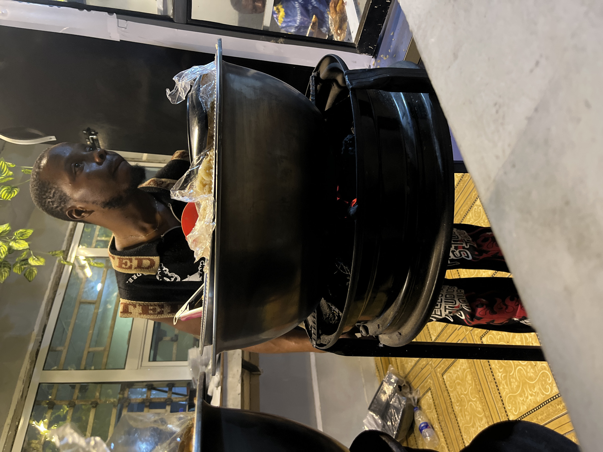 The image depicts an outdoor food preparation scene, likely a street food vendor, in what is plausibly Kubwa, Nigeria. A man with short hair and dark skin is positioned behind a large, black, multi-tiered cooking pot or roasting equipment. The pot contains apparent fuel/fire in the base section with a cooking area at the top. Food (potentially dough-like items) is partially visible, some wrapped in a clear plastic bag. He is wearing a black shirt with tan/brown accents along the collar and shoulder. Behind him, through what seems to be a building window, one can see plants and a window with metal bars. The floor in the foreground is covered in yellow tiles. A water bottle is visible on the floor. The environment suggests an urban or semi-urban outdoor setting.
