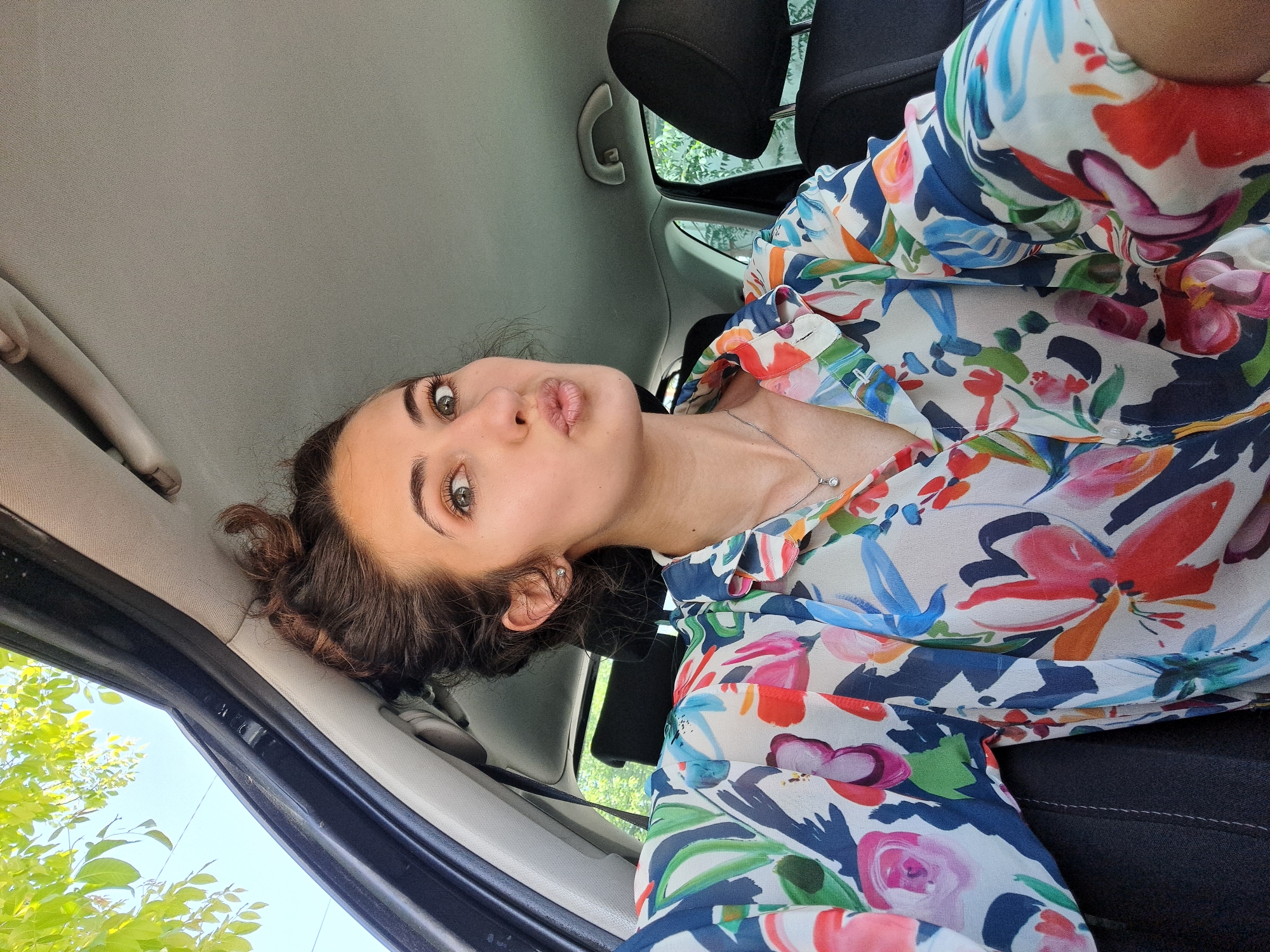 The image is a selfie taken from inside a car. The subject is a woman with brown hair and green eyes. She is wearing a colorful floral-patterned blouse and a silver necklace. Her hair is styled in a loose bun. She has her lips puckered as if blowing a kiss. The car's interior is visible, including the headrest, ceiling, and door frame. Outside the window, green trees are visible. There are no identifying features to suggest the location in Sofia, Bulgaria.
