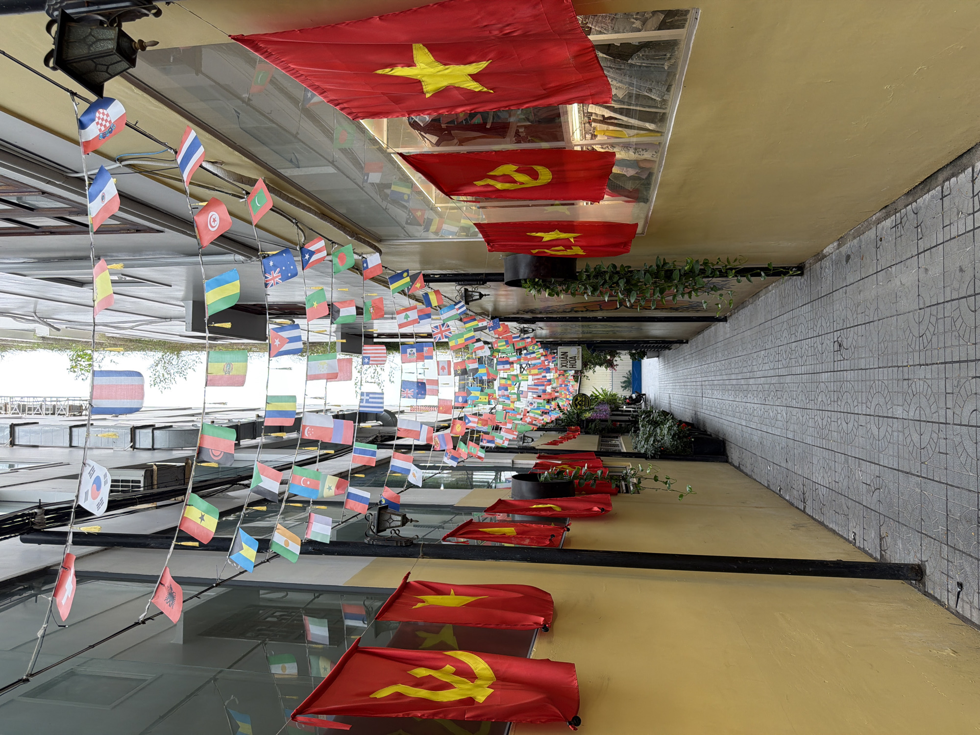 The image shows an outdoor alleyway in Nha Trang Ward, Vietnam, decorated with numerous national flags hanging overhead. At the far end, there are hanging plants above a passageway with a few people partially visible in the distance. The near end features a series of larger Vietnamese flags interspersed with flags displaying a communist hammer and sickle symbol. The right side of the image features a wall covered with grey tiles, while the opposite side reflects buildings and sky. The surface below the hanging flags is painted a yellow color.