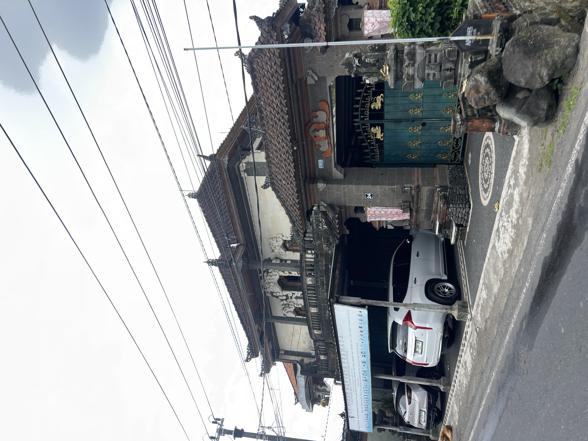 The image shows a two-story building with traditional Balinese architecture in Abiansemal, Indonesia. The building has a dark tiled roof with ornate carvings. The facade features intricate stone details, balconies, and windows. In front of the building, there is a gated entrance with a teal-colored door adorned with gold lion motifs. To the left of the entrance, a banner with text and various icons is displayed. Two white cars are parked under a covered area in front of the banner. A small dog is visible lying on the ground to the left. The street in front of the property is paved, and there are large rocks placed along the side of the road. The sky is overcast.