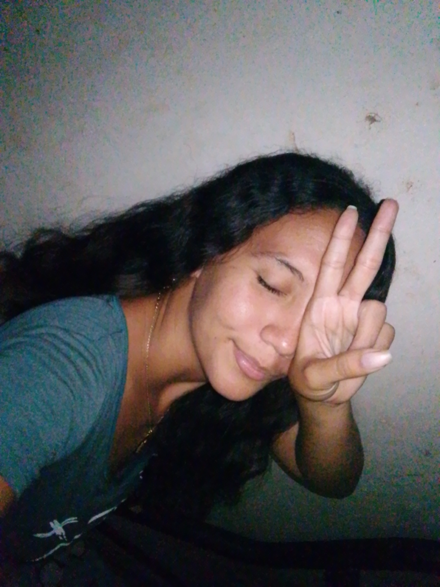 A dark-haired woman poses indoors against a light-colored wall. She is wearing a t-shirt and a necklace. She has her eyes closed and is holding up two fingers in a peace sign over her face. The location of the image is unknown and cannot be assumed as Juan Griego, Venezuela.