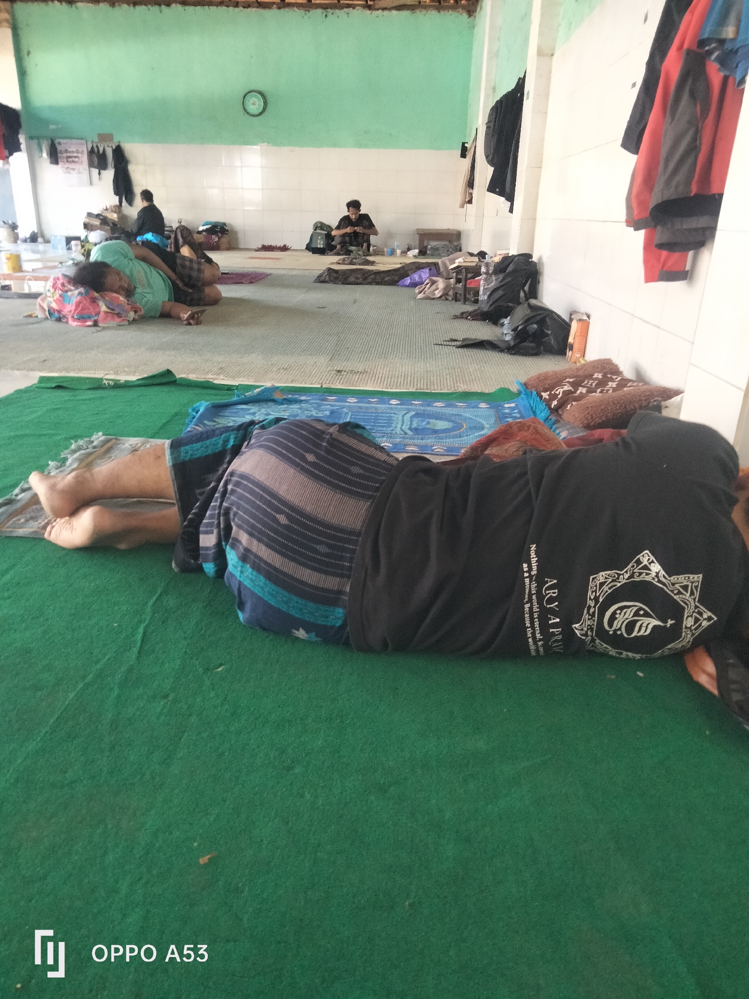The image shows the interior of a room with green carpeting and light-colored tiled walls. The room appears to be a communal sleeping area, possibly a mosque or shelter, given the presence of prayer mats and people resting on the floor. In the foreground, a person is lying on their side on a green carpet, covered by a patterned cloth and wearing a black shirt with white text and a circular emblem. Their legs are visible, with bare feet resting on a prayer mat. In the background, other individuals are also resting on the floor. One person in a light blue shirt is lying on a mat, while another person in a dark shirt sits on the floor facing away from the camera. Various bags and belongings are scattered around the room, and clothing items are hung on the wall. A clock is visible on the green wall in the background. The overall scene depicts a quiet, communal space where people are resting or sleeping. The location is identified as Badung, Indonesia.