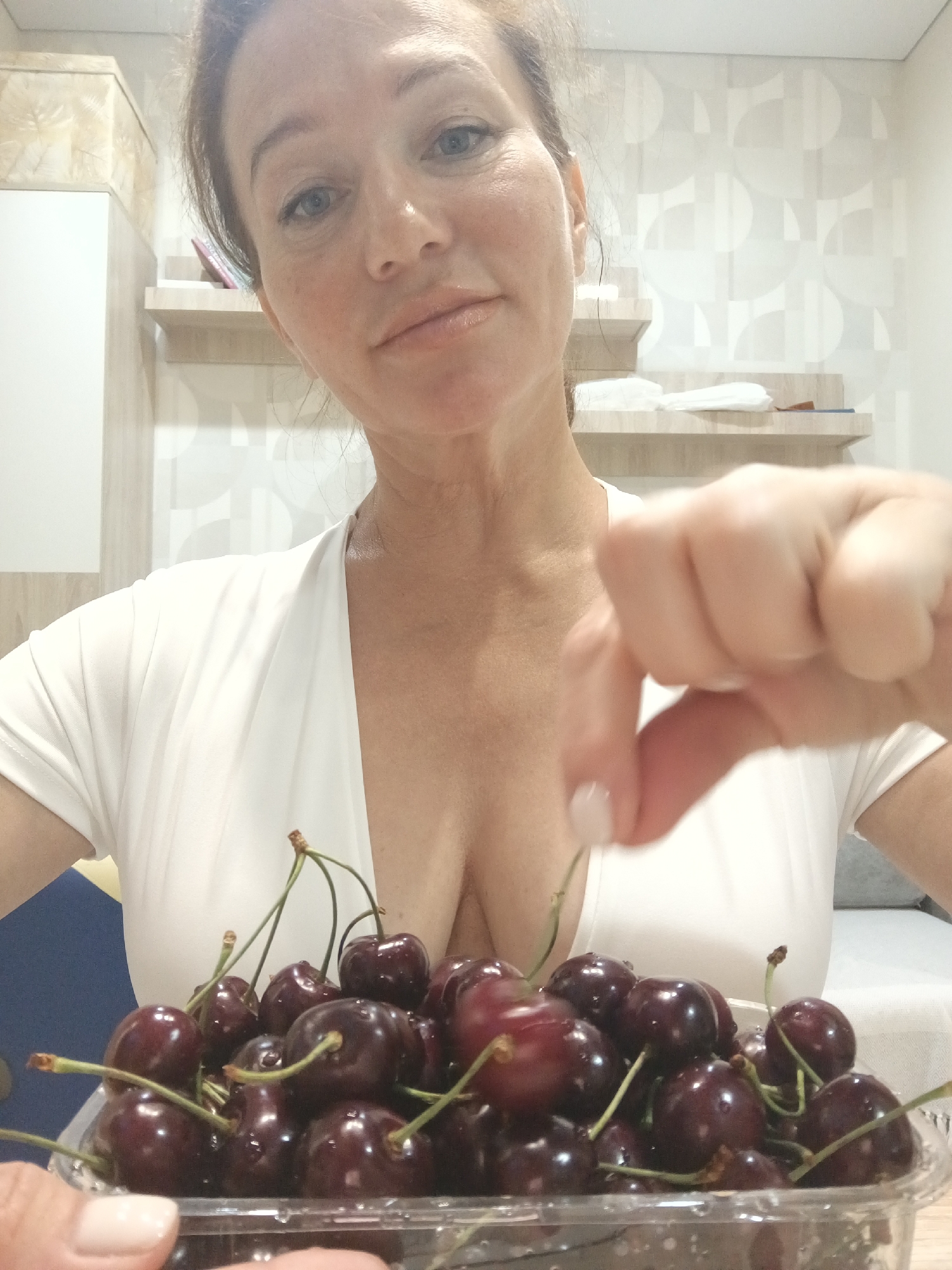 A woman is taking a selfie while holding a clear plastic container of dark red cherries. She is wearing a white V-neck top. Her right hand is extended towards the viewer, with her index finger and thumb pinching a cherry stem. The cherries are glistening with water droplets. In the background, there are shelves with some items on them, and a patterned wallpaper. The image does not provide any information to determine the location in Sochi, Russia.