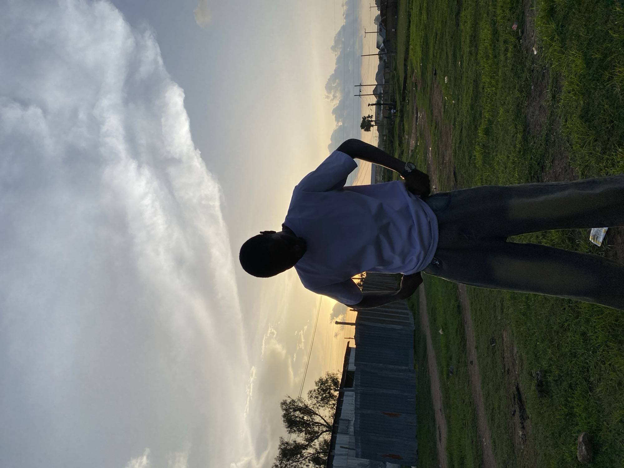 The image depicts a male figure standing outdoors in Bukuru, Nigeria, during either sunrise or sunset, indicated by the strong backlighting. The figure, positioned slightly right of center, is silhouetted against the bright horizon, facing forward with his head tilted downwards. He wears a light-colored short-sleeved t-shirt and dark, wide-leg pants, with his hands placed on his hips. A watch is visible on his left wrist.
The upper portion of the frame is dominated by a sky displaying warm orange, yellow, and peach hues near the horizon, transitioning to light blue and grey tones upwards. Scattered clouds, varying from wispy to more dense formations, are visible.
In the mid-ground, behind the person on the left, are structures constructed from corrugated metal sheets, resembling fences or makeshift buildings, partially obscured by sparse trees. Utility poles and overhead lines are present across the mid-horizon. On the right side, the landscape opens to reveal more distant, low-rise structures, a single palm tree, and additional utility poles and lines. The ground is a mix of green grass, dirt paths, and sparse vegetation, with scattered debris visible in the foreground.