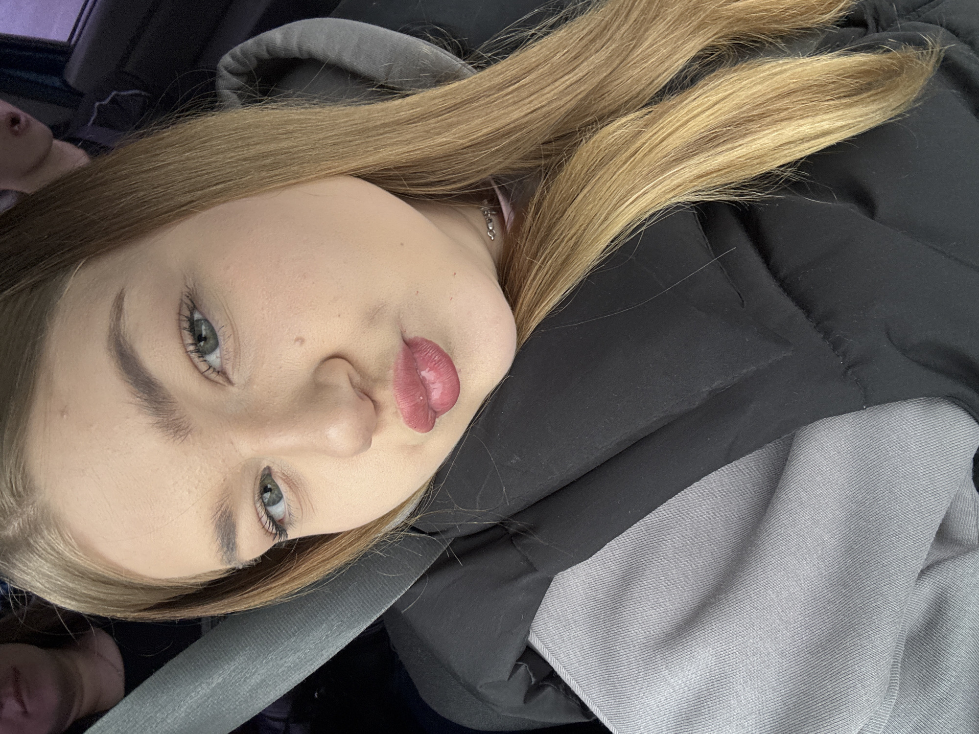 A young woman with blonde hair and light eyes is pictured indoors, seemingly in a vehicle. She is wearing a dark jacket and a grey hoodie, secured by a seatbelt. The location is unknown, and there is no indication the picture was taken in Chaikovsky, Russia.