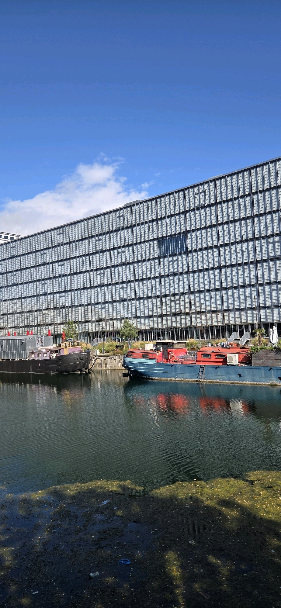 The image depicts a modern, multi-story building with a facade composed of numerous glass panels, reflecting the blue sky and scattered clouds. In the foreground, a body of water, possibly a canal or river, is visible. Two boats are moored on the water; one is a dark-colored barge with purple accents, and the other is a red and blue boat with a cabin. The water surface exhibits ripples, and the lower portion of the image shows debris floating on or near the water's edge, including what appears to be plastic waste and organic matter. Trees and some vegetation are present along the bank behind the boats and in front of the building. The scene appears to be an urban waterfront environment, likely a commercial or residential area given the building's architecture. This image is not from Strasbourg, France.