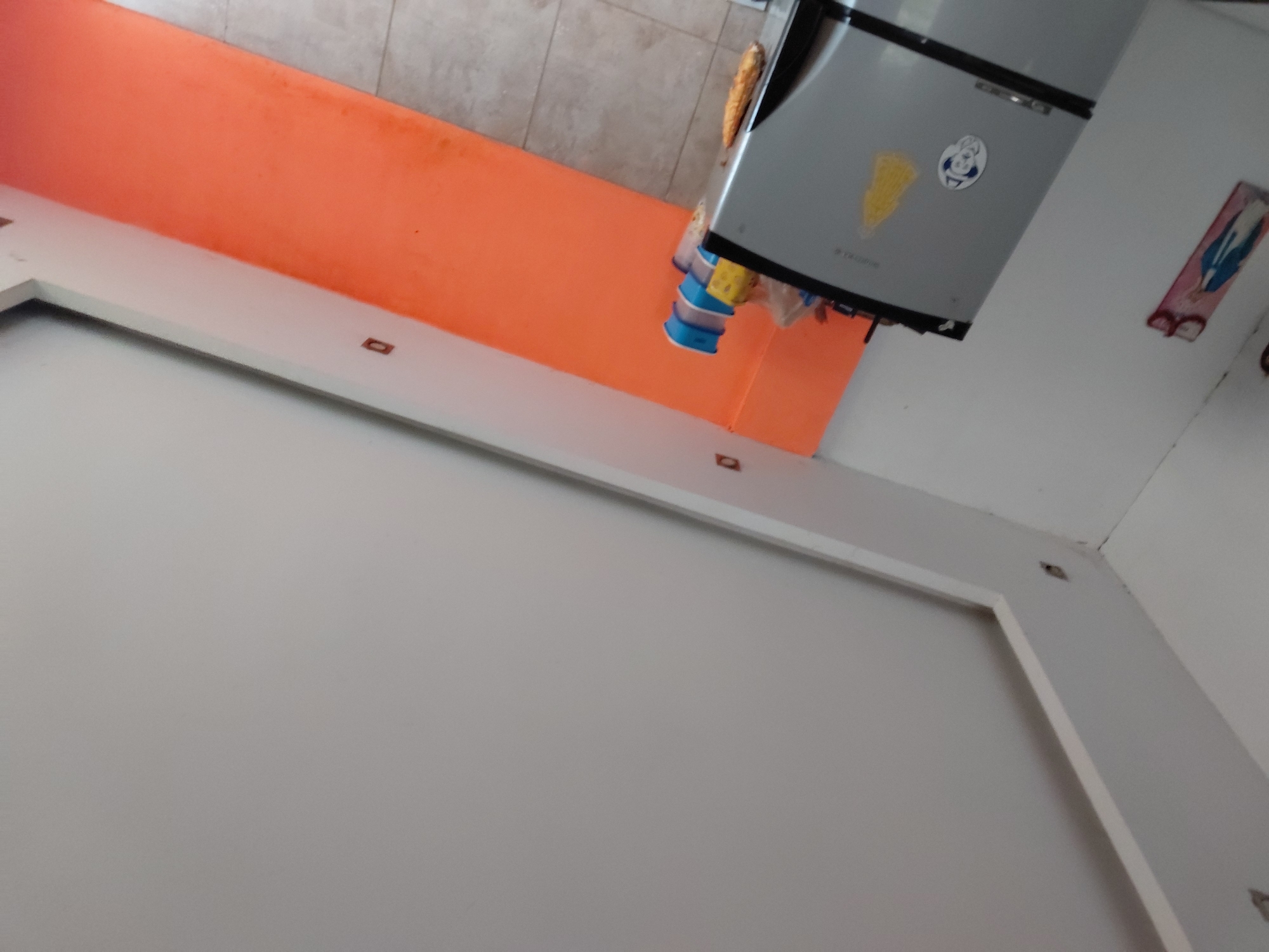 The image displays an interior scene, captured with a strong counter-clockwise tilt, suggesting a view looking upwards from a low angle. When viewed upright, the foreground reveals a light grey wall or floor framed by a thick, white, rectangular border. Several small, square-shaped indentations or fixtures are visible within this white border and on the surface beyond.
An orange-colored surface, likely a wall or counter, stretches horizontally across the mid-ground. Above this orange section, a portion of a light-toned, possibly tiled, wall is visible.
A prominent silver-grey refrigerator occupies the upper right side of the frame. Its front door features decorative elements, including a yellow, triangular sticker or magnet and a white, circular one with a blue outline of a figure or logo. Below these, faint, dark text, possibly branding, is visible but illegible. Beneath the refrigerator, a cluster of blue plastic cups appears to be stacked or hanging, alongside other miscellaneous items, possibly in plastic bags.
To the right of the refrigerator, on an adjacent white wall, a rectangular piece of artwork or decoration is mounted, featuring blue, pink, and white elements. Below this, a pair of eyeglasses is hanging. The scene is static, devoid of people or specific activities, depicting what appears to be a functional domestic space. No direct sources of natural light are visible, making the time of day indeterminate.