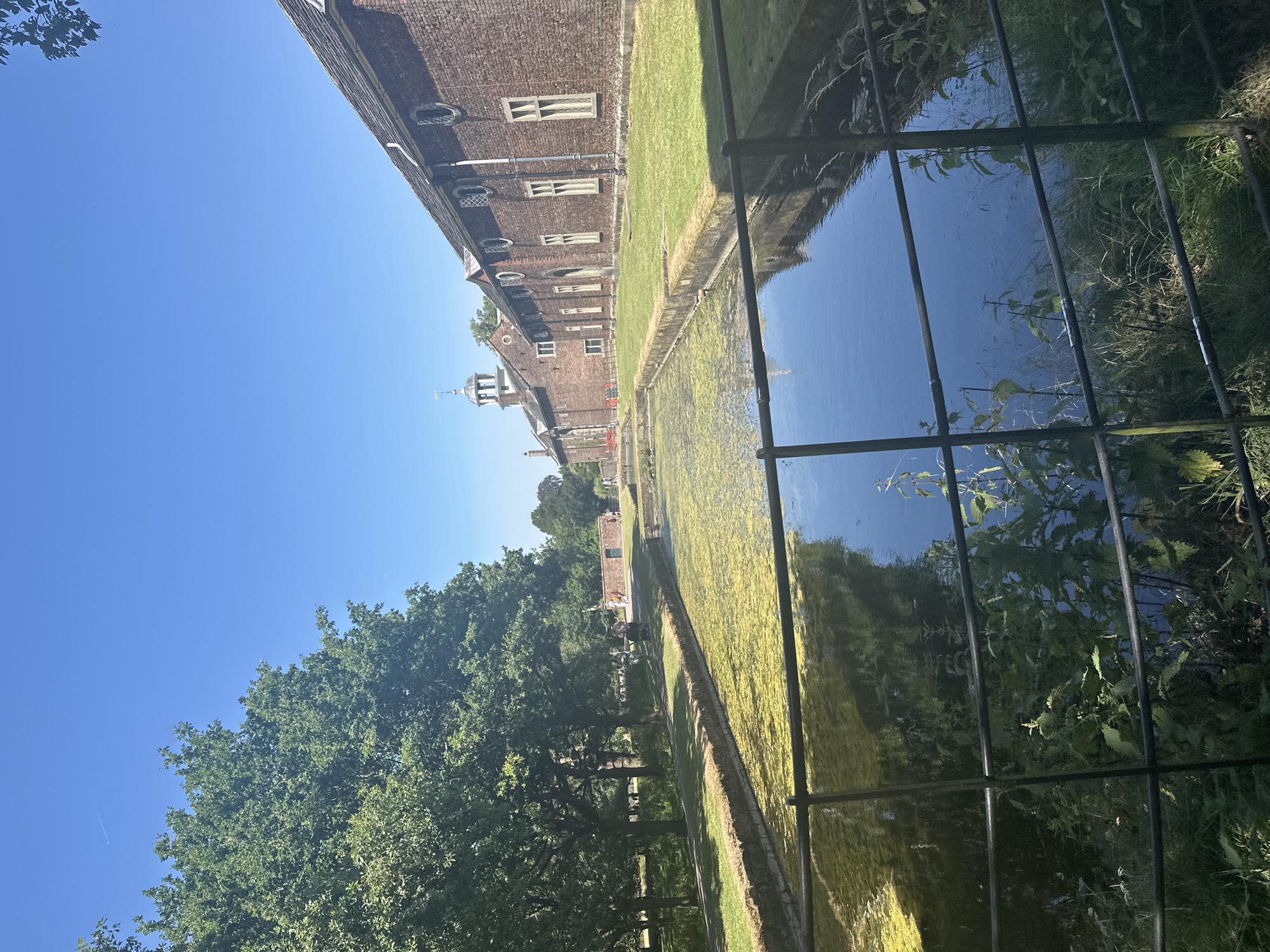 This image captures a canal-like body of water, partially covered in green algae, with a dark metal fence in the foreground. On the right side of the water, a long, red brick building with a tiled roof runs parallel to the canal. This building features numerous windows with light-colored frames. Further down the canal, a smaller brick structure with a pitched roof is visible. On the left side of the canal, a grassy area is lined with mature trees, and beyond that, more greenery and distant buildings can be seen. The sky is clear and blue. The location is identified as being in the city of Trafford, United Kingdom.