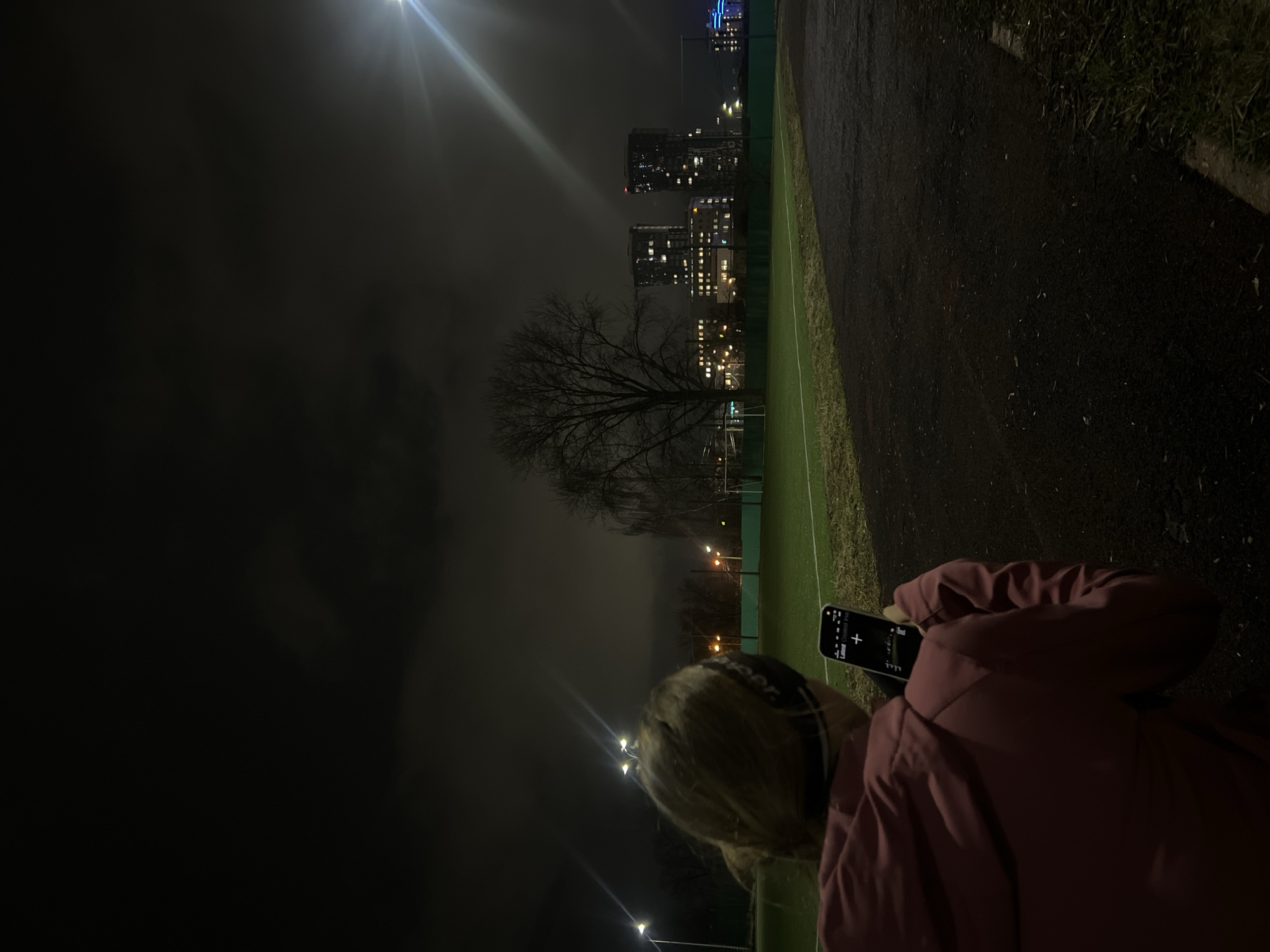 A nighttime photograph taken in Kazan, Russia, shows a person facing left, partially visible from the back. The person, wearing headphones and a maroon jacket, is looking at a smartphone. The background includes a park with a soccer field, bare trees, street lights, buildings in the distance, and an overcast sky. The location is illuminated by artificial lighting.
