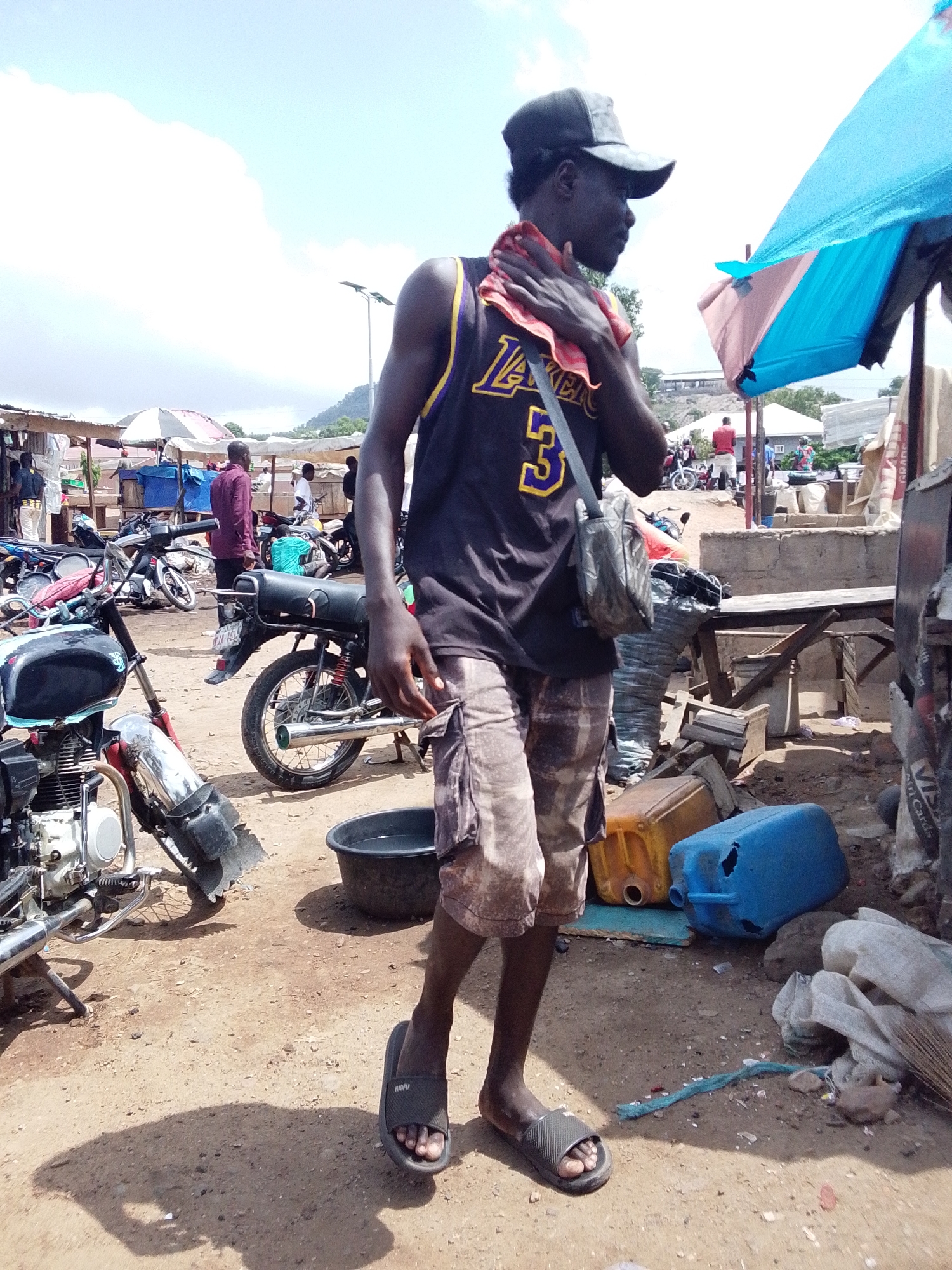 The image shows a man in Lokoja, Nigeria. He is wearing a black Lakers jersey with the number 3, patterned shorts, and black sandals. He has a black cap on his head and a bag slung over his shoulder. He is holding a red cloth to his neck.
The background shows a bustling outdoor scene with motorcycles, vendor stalls, and other people. There are blue and orange plastic containers, some trash, and a crude wooden bench nearby. The ground is dirt.