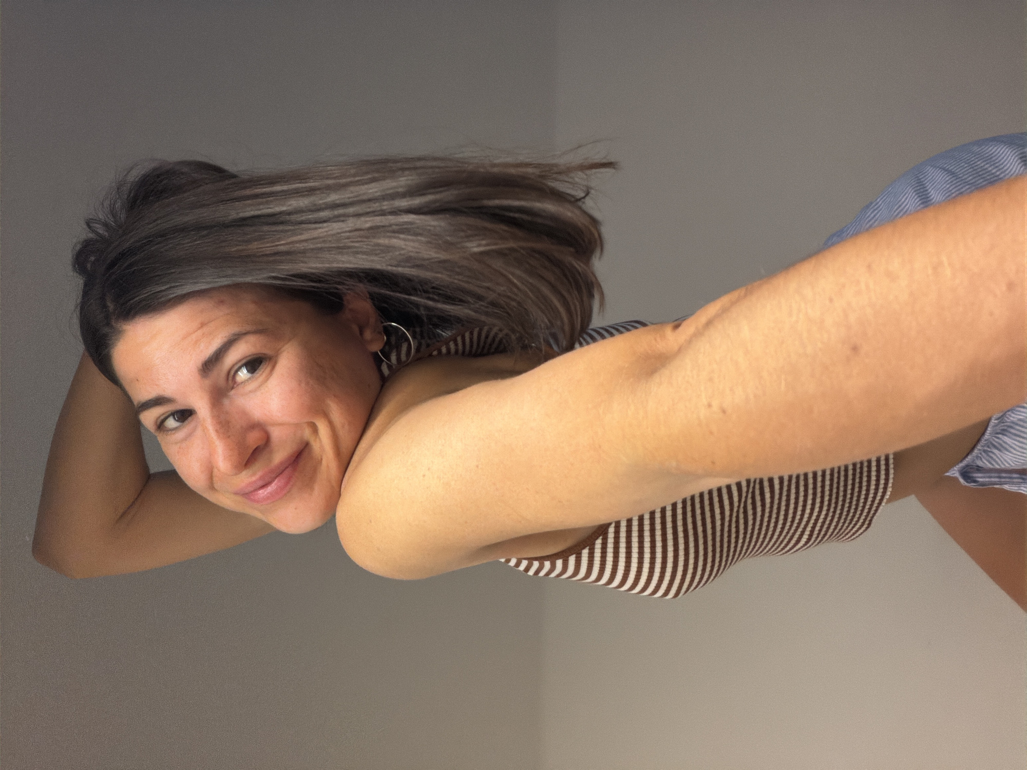 A woman with brown, shoulder-length hair and light skin is smiling at the camera. She is wearing a brown and white striped top and blue striped shorts. She has a hoop earring in her left ear. Her left arm is raised and bent behind her head. The background is a plain, light grey wall. The location is not discernible from the image.