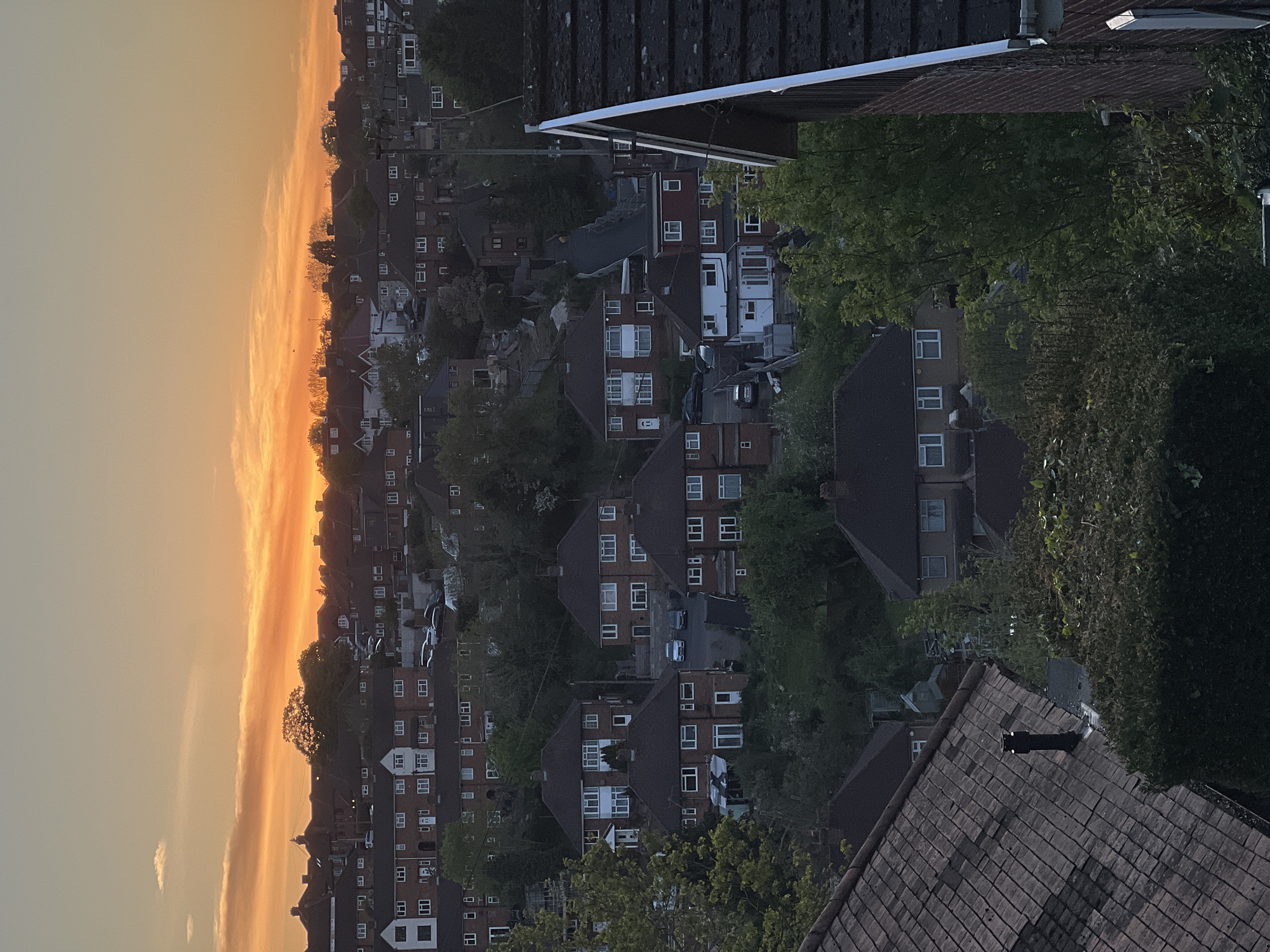 The image, rotated 90 degrees clockwise, presents a panoramic view of a residential area in High Wycombe, United Kingdom, under the warm glow of a sunset.
In the immediate foreground, at the bottom of the correctly oriented image, a large, dark-tiled roof with a visible chimney or vent occupies the lower section, indicating the viewpoint is from an elevated position, possibly another building. Further into the scene, a dense arrangement of suburban houses extends across the frame. These are predominantly two-story, brick buildings with pitched, dark-tiled roofs and multiple windows, often featuring contrasting white frames. The architectural style is typical of British housing.
Numerous mature trees with dark green foliage are interspersed among the houses, partially obscuring some dwellings and adding a natural element to the urban landscape. Small glimpses of parked cars are visible on driveways or narrow streets between the houses.
The right side of the image, when correctly oriented, is dominated by a vibrant sky, showcasing the intense orange and yellow hues of a sunset. Streaks of thin clouds catch the bright light, stretching horizontally across the horizon, contrasting with the paler orange and grey tones of the upper sky. The light on the houses and trees is dimming, indicating the end of the day.
No specific human activity is discernible within the frame. There is no legible text visible on any buildings, vehicles, or signs.