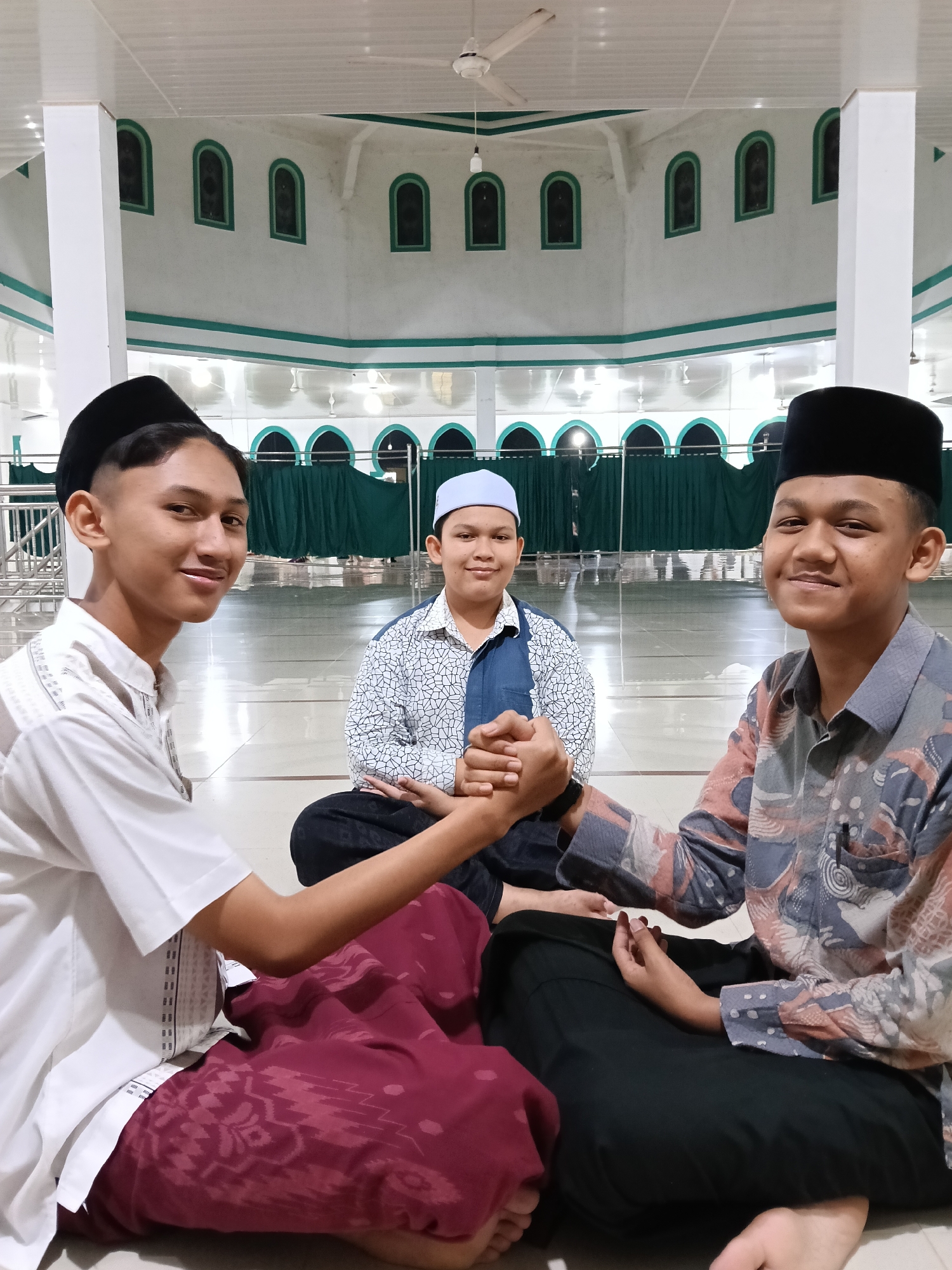 Three young men are seated on the floor of what appears to be a mosque. The two men on the left and right are shaking hands. The man on the left is wearing a white shirt and maroon sarong, while the man on the right is wearing a patterned shirt and dark pants. Both are wearing black peci hats. The man in the middle is wearing a patterned shirt with a blue vest and a white peci. The background shows a large room with white walls, green trim, arched windows, and green curtains. The location is likely a mosque in Lhokseumawe, Indonesia.