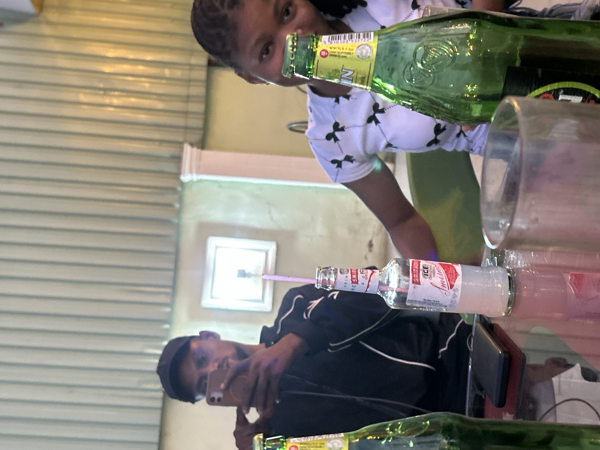 The image depicts a casual indoor scene at an establishment in an unidentified location in Unknown, Unknown. Two individuals are visible, along with various objects on a table.
A woman with braided hair, wearing a white top with black bow patterns, is positioned in the upper part of the frame. She is holding a green glass bottle.
Below her, a man wearing a black baseball cap and a black and white jacket is holding a smartphone, presumably taking a photo or video.
The table contains a partially visible glass bottle, a bottle of what appears to be Smirnoff Ice with a straw, a glass, and a smartphone.
The environment includes a wall with horizontal metal panels and a window with a frame. A partially visible column is also present.