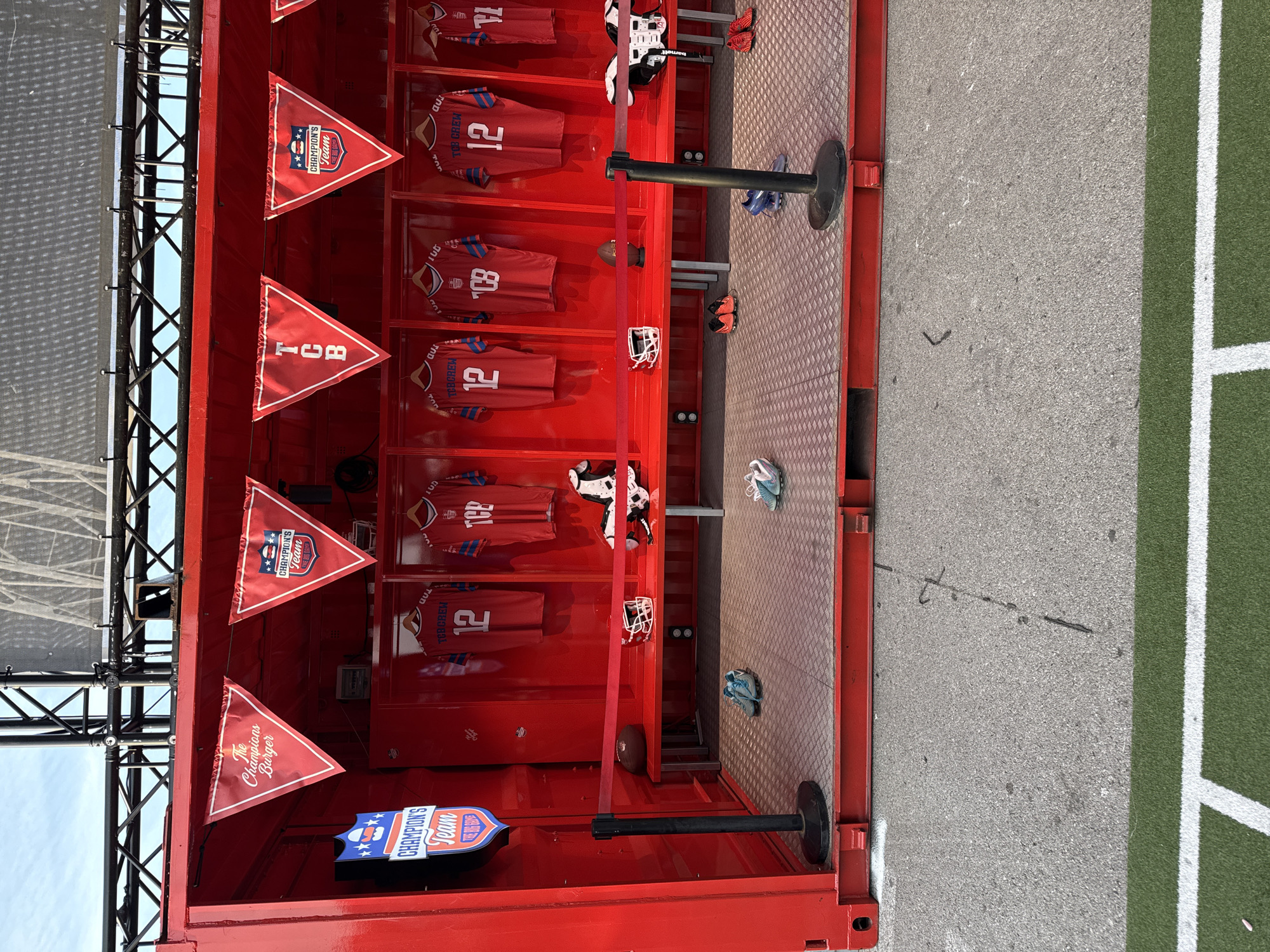 The image depicts the interior of a red container or booth, decorated with sports-themed items. Several red jerseys with the number "12" and "TCB CREW" printed on them are displayed on hangers. Football helmets and shoes are also present. Triangular flags with "TCB" and "CHAMPION'S Team" printed on them hang from the top of the container. The floor of the container is made of a textured, possibly metal, material. Outside the container, there is a section of pavement and a green artificial turf resembling a sports field. The image provides no information about the location in Barcelona, Spain.