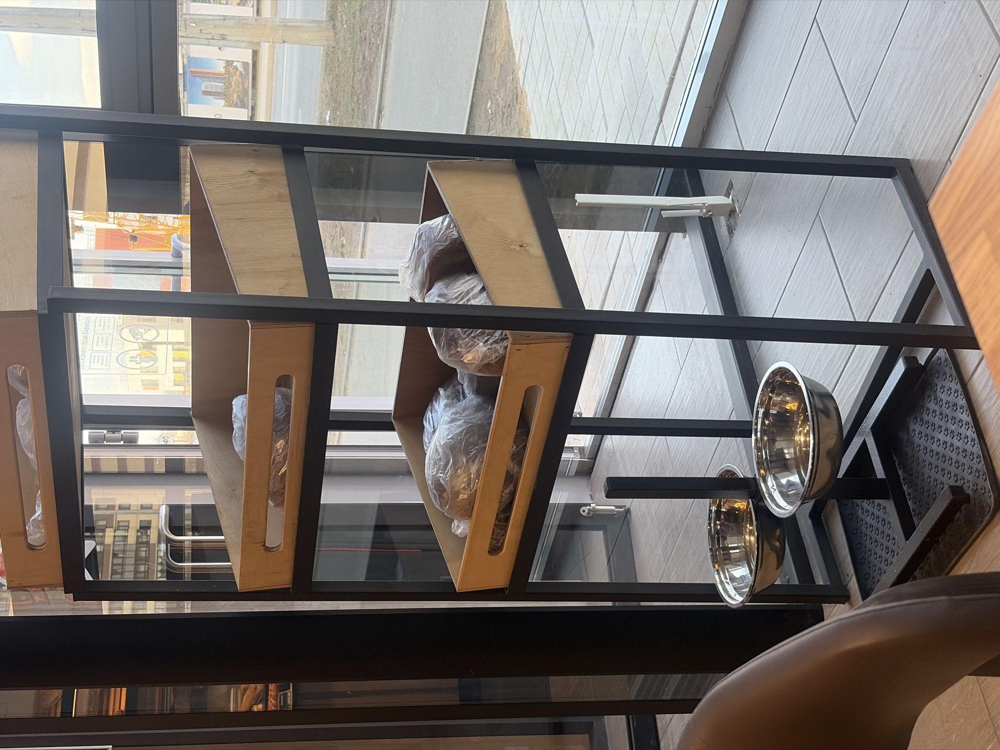 The image depicts the interior of a commercial establishment, likely a bakery or cafe, viewed towards large street-facing windows in Saint Petersburg, Russia. The specific location is strongly indicated by a billboard visible through the window, which features modern residential high-rise buildings resembling the LEGENDA Heroes Park complex, placing the scene in the vicinity of **Prospekt Geroyev, Krasnoselsky District, Saint Petersburg**.
In the foreground and center, a black metal shelving unit with three levels is prominent. Each level holds a light-colored wooden display crate. The middle and bottom crates contain multiple items, appearing to be loaves of bread or similar baked goods, individually wrapped in clear plastic bags. The top crate also contains wrapped items, partially visible.
Below the shelving unit, two polished stainless steel pet bowls are affixed, positioned directly above a black mat embossed with paw print patterns. This indicates the establishment is pet-friendly. A section of a dark brown chair, likely customer seating, is visible in the lower left corner. The floor consists of light-colored rectangular tiles.
Through the expansive windows, an urban exterior is visible. A paved sidewalk or patio transitions to a street with a utility pole on the right. In the middle-distance background, a large yellow construction crane signifies ongoing development. To the right, a billboard displays the aforementioned modern residential buildings. Reflections in the window show Russian text, "ДВЕРЬ ОТКРЫВАЕТСЯ НА СЕБЯ" (The door opens towards you), and a white sign with black icons depicting a surveillance camera and a paw print, indicating CCTV and pet accommodation. No individuals are present in the visible scene.