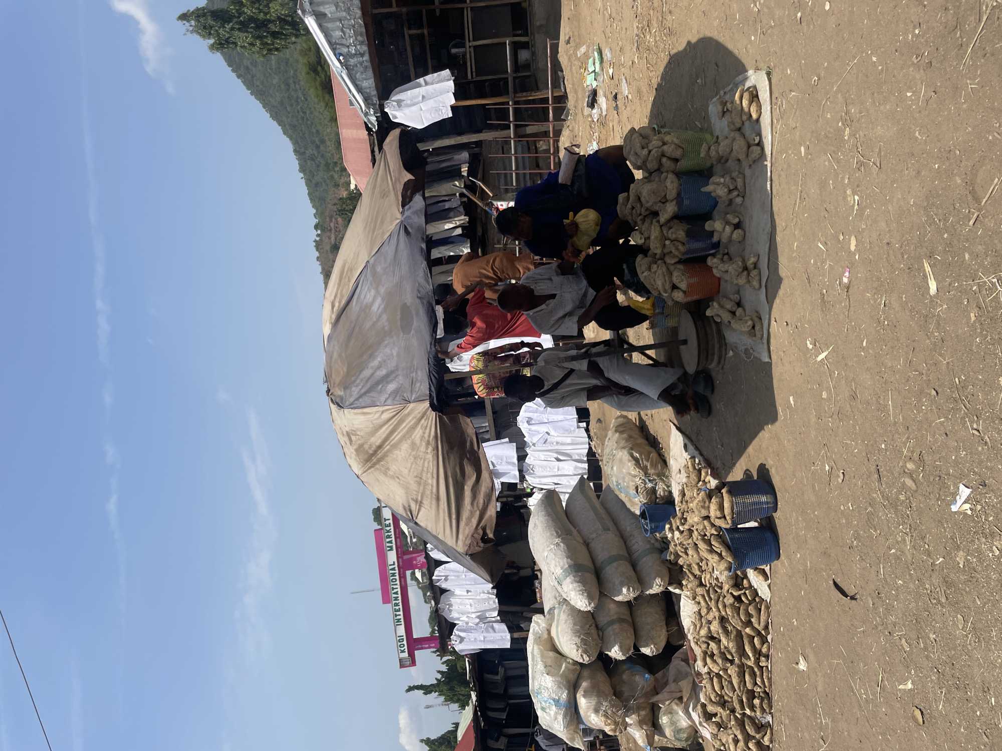 The image depicts an outdoor market scene under a clear blue sky, likely during daytime given the bright sunlight and visible shadows. The ground is unpaved, dusty, and uneven.
In the foreground and midground, numerous people are gathered around various market stalls. A large, beige-colored umbrella provides shade over a section where several individuals are seated. Some people are interacting with the goods on display, possibly engaged in buying or selling. One person in a blue top is seen handling items, while others are dressed in various casual attires, including a prominent figure in a white top.
The primary merchandise visible consists of a large quantity of root vegetables, likely yams, which are arranged on tarps on the ground, packed into colorful woven baskets (blue, green, orange), and stored in large white sacks. Further back, more white shirts are seen hanging, suggesting another section of the market.
In the background, lush green mountains or hills rise under the bright sky, adding to the outdoor setting. Simple market structures, possibly constructed from wood and corrugated metal, are also visible. A prominent pink sign with vertical text reads "KOBI INTERNATIONAL MARKET," providing a specific location identifier for this bustling commercial area.