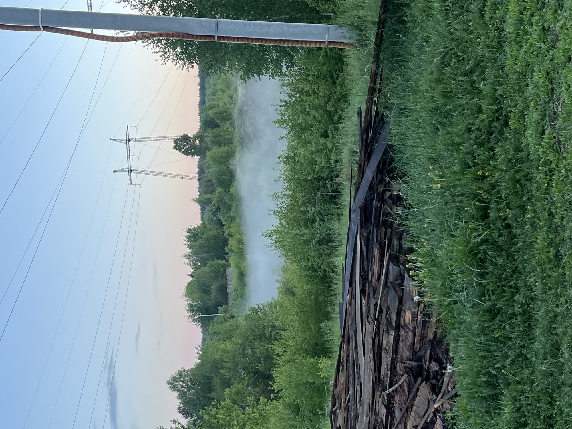The image depicts a landscape in what appears to be a rural or semi-rural area near Nizhny Tagil, Russia. In the distance are power lines stretching across a hazy or foggy body of water, possibly a river or lake, bordered by dense vegetation. The fog obscures part of the waterway and the treeline. Closer to the foreground is a pile of weathered wooden planks, possibly from an old fence or building. To the right of the planks, there's lush, green grass and foliage. A utility pole and associated wires are visible in the top left corner. The sky is a light blue with a hint of pink or orange near the horizon, suggesting either dawn or dusk. There are no visible people in the image.