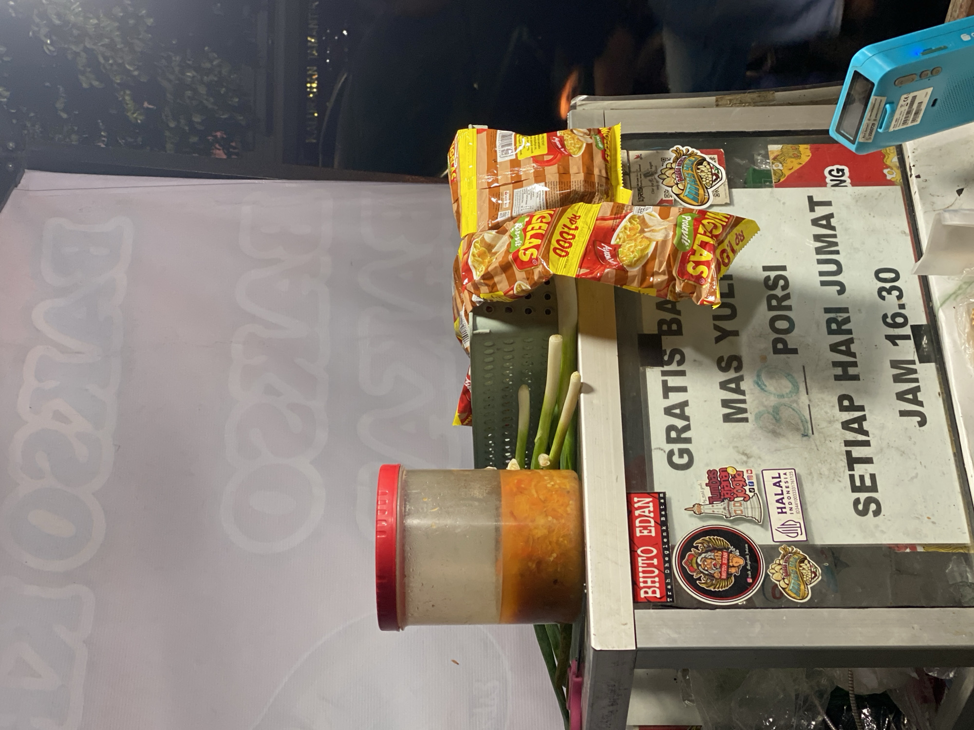 The image shows a food stall, likely selling bakso, in Kasihan, Indonesia. The stall has a white banner with the word "BAKSO" written in large, faded letters. On the front of the stall, there is a clear container with red sauce or chili paste, surrounded by green onions. Several packets of instant noodles are hanging from the stall. Stickers adorn the front, including one with "BHUTO EDAN TRAH DHEGLENK BATAM" and a "HALAL INDONESIA" logo. A prominent sign reads "GRATIS MAS YULI 30 PORSI SETIAP HARI JUMAT JAM 16.30", indicating a promotion for free portions every Friday at 4:30 PM. A bright blue electronic device, possibly a payment terminal, is visible in the lower right corner. The background suggests an outdoor setting at night, with trees and building lights visible.