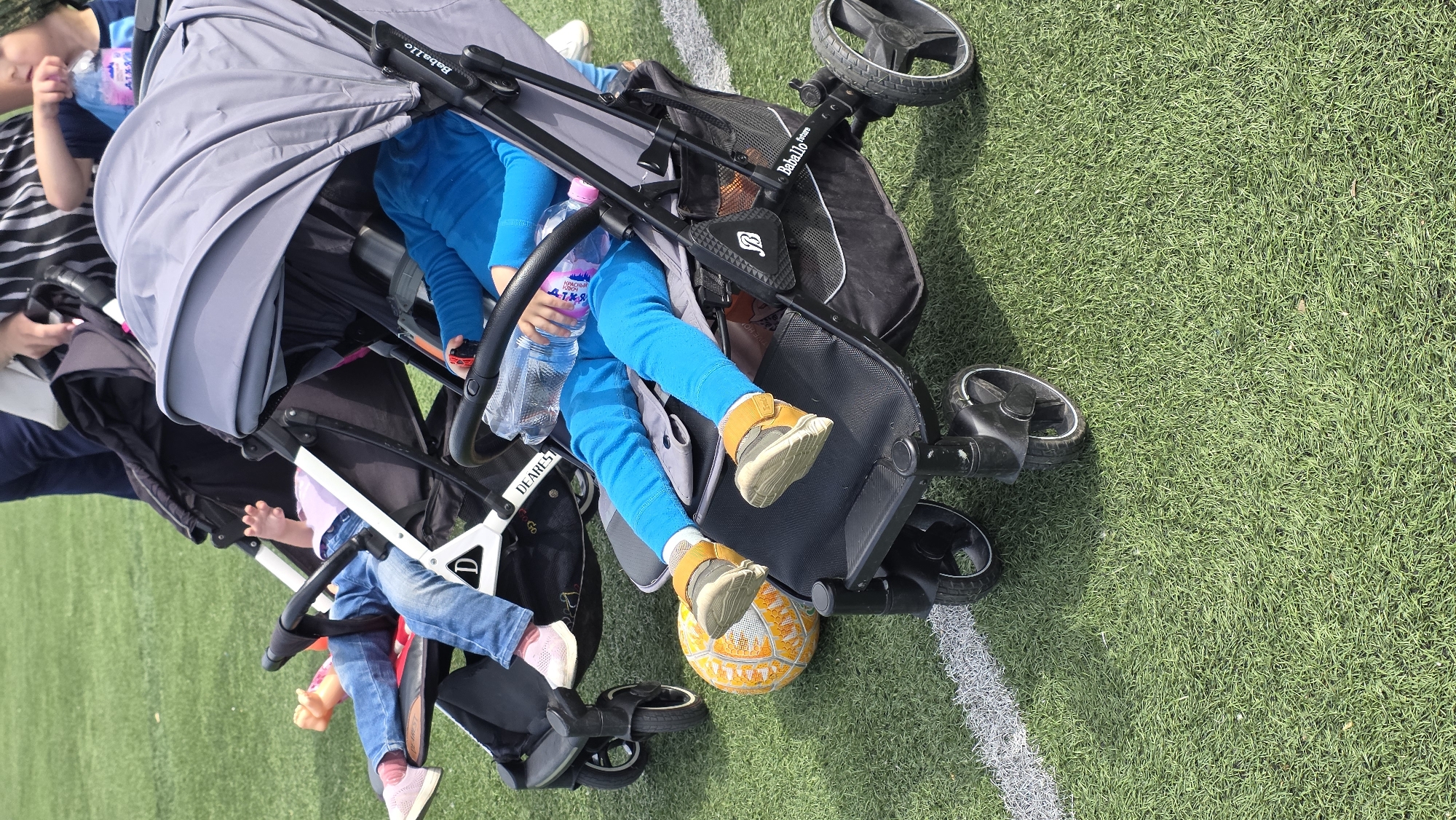 The image captures a scene on a vibrant green, synthetic turf field marked with a crisp white boundary line. Two strollers are centrally positioned, each occupied by a child. The child in the top stroller is clad in bright blue attire, holding a water bottle, with their feet resting on a yellow-orange soccer ball. The second child in the bottom stroller is dressed in jeans, with pink sneakers. To the left, an adult is partially visible, wearing a striped shirt. The location is in an unknown city.