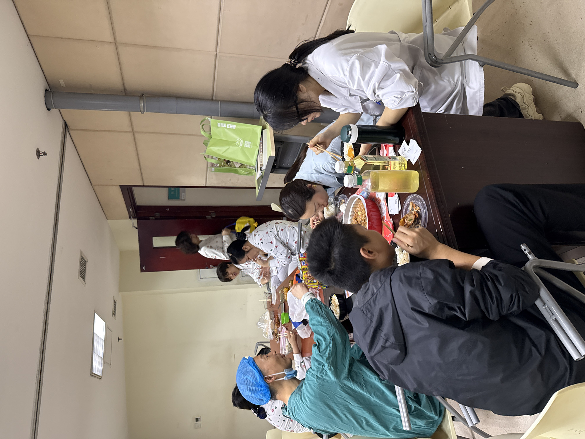 This image, rotated 90 degrees counter-clockwise, depicts a group of people having a communal meal in what appears to be an institutional or educational setting.
Approximately eight individuals are seated around two long tables, engaged in eating. The attire of the people suggests a connection to the medical field: one person in the foreground (far left when upright) wears teal medical scrubs and a blue surgical cap, while others are dressed in white lab coats or patterned white tops that resemble hospital gowns or pajamas. Most are young adults, focused on their food and using chopsticks.
The tables are laden with numerous plastic containers filled with various dishes, including rice, stir-fries, and other Asian cuisine, along with plastic bottles of beverages and a thermos. The setting is functional, featuring light-colored walls, standard ceiling tiles, and fluorescent lighting. A prominent grey pipe runs along the upper wall/ceiling junction, and multiple ventilation grates are visible on the walls. A fire sprinkler head is also visible on the wall in the upper left section of the image (when upright). A dark wooden door is partially visible in the background, along with a light switch or thermostat on a wall.
A notable detail is a green reusable shopping bag placed on a surface next to one of the diners, which clearly displays the text "重庆医科大学" (Chóngqìng Yīkē Dàxué), identifying the location as Chongqing Medical University in Yuzhong District, China. This confirms the scene likely takes place within a university dormitory, cafeteria, or a break room for students or staff. The atmosphere appears relaxed, typical of a mealtime break.