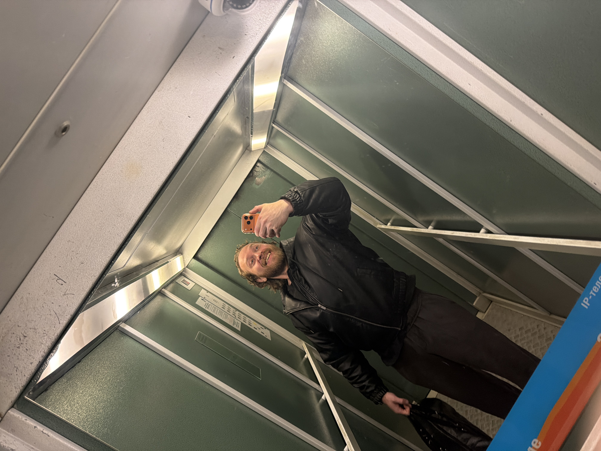 A man is taking a selfie in an elevator. He is holding an orange phone. He has curly hair and a beard. He is wearing a black jacket and dark pants, and holding a black bag. The elevator is paneled with green. There is a security camera visible in the top left corner of the frame. There are white lines on the elevator walls. The location is Saint Petersburg, Russia.
