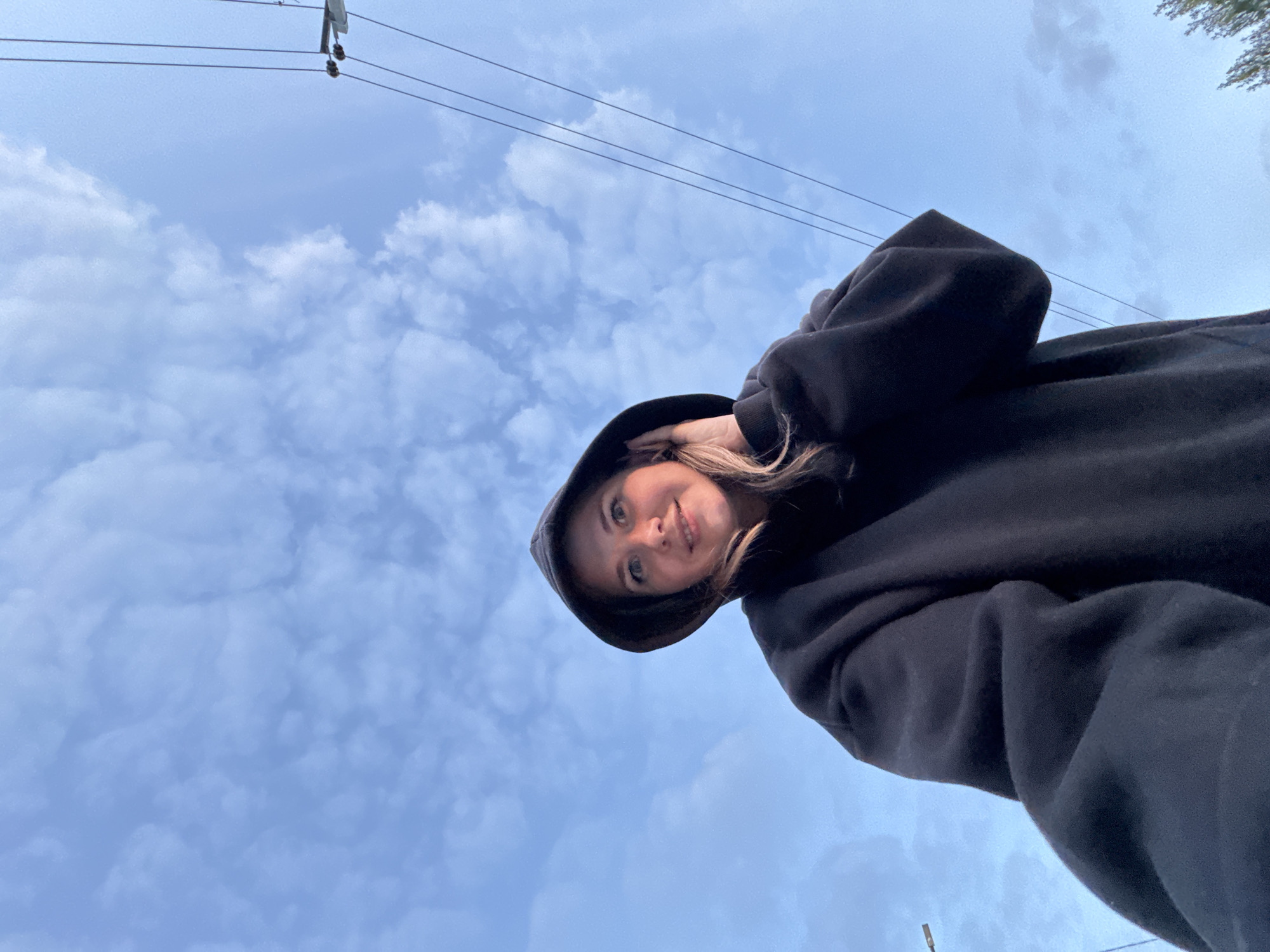 A low-angle shot shows a young woman wearing a black hoodie standing outdoors. She is centered to the right of the frame. The sky, filled with puffy white clouds, occupies most of the background. Some utility lines run across the top of the frame. The location is in Горный, Russia.
