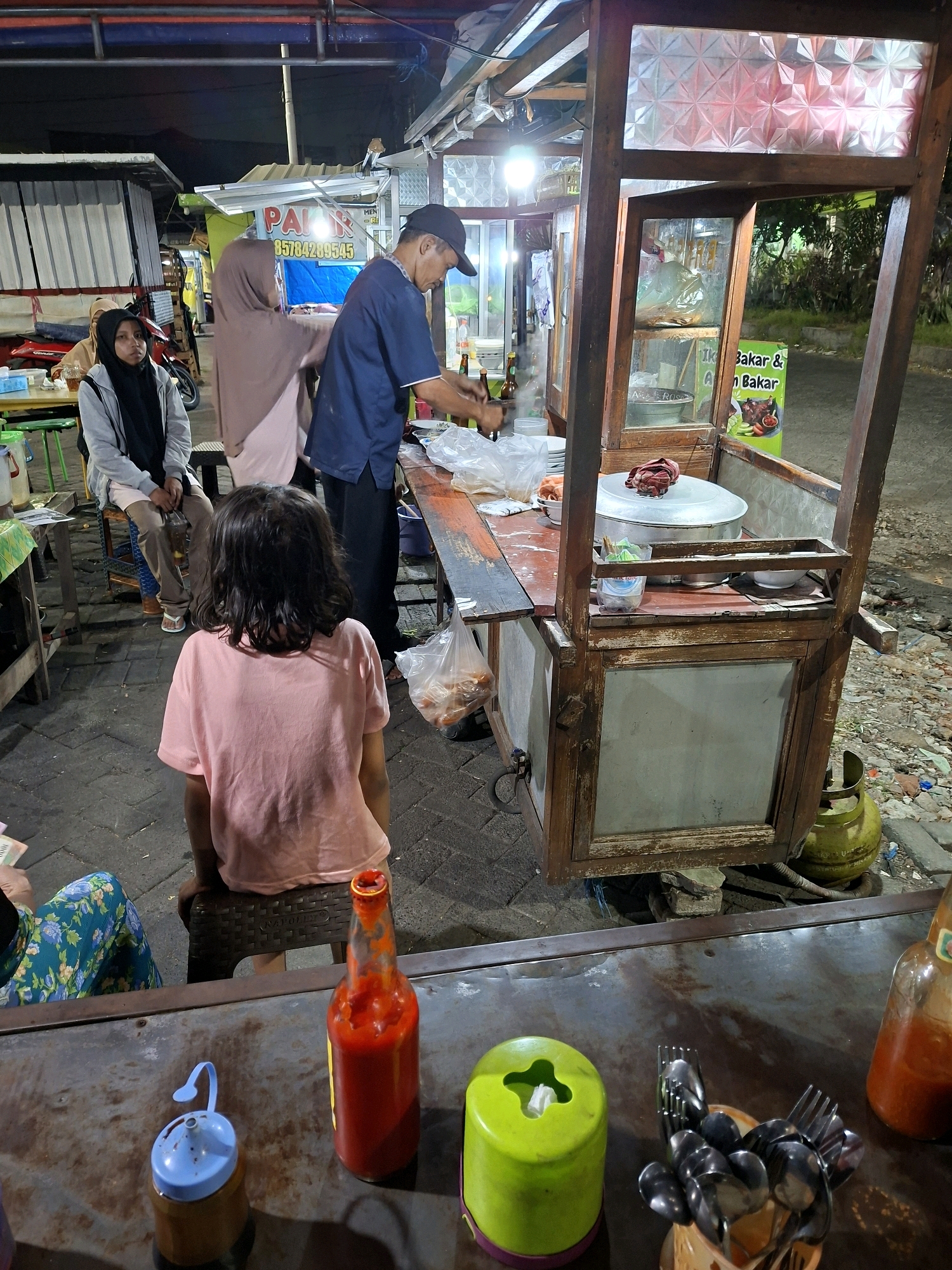 The image depicts an outdoor food stall, likely in Surabaya, Indonesia, at night. A man in a blue shirt and a black cap is preparing food behind a wooden cart. To his left, a woman wearing a hijab and a grey jacket is seated on a stool, looking towards the food stall. A young girl in a pink t-shirt is also seated, facing away from the camera, with her back towards the food preparation area. In the foreground, a metal table holds various items, including a red bottle of sauce, a blue dispenser, a green tissue holder, and a collection of cutlery. The background shows more stalls and vehicles, with indications of a street or pathway. The lighting suggests an urban environment, possibly a night market or a street food vendor area. A gas cylinder is visible to the right of the food cart.