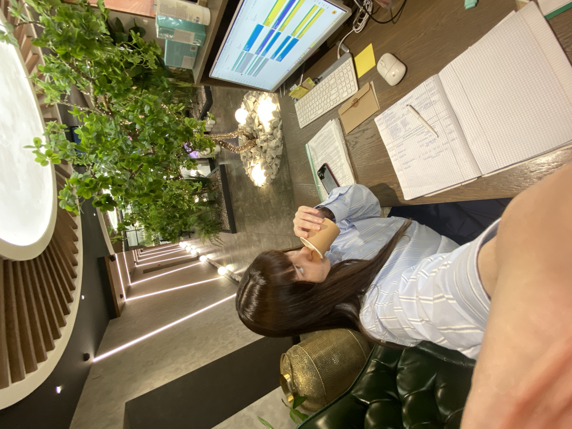 A woman in a light blue striped shirt sits at a desk, drinking from a disposable cup. The desk has a computer monitor displaying a chart, a keyboard, a mouse, a notebook with writing, and a pen. The surroundings feature modern architecture with indirect lighting and decorative plants. The location is unknown.