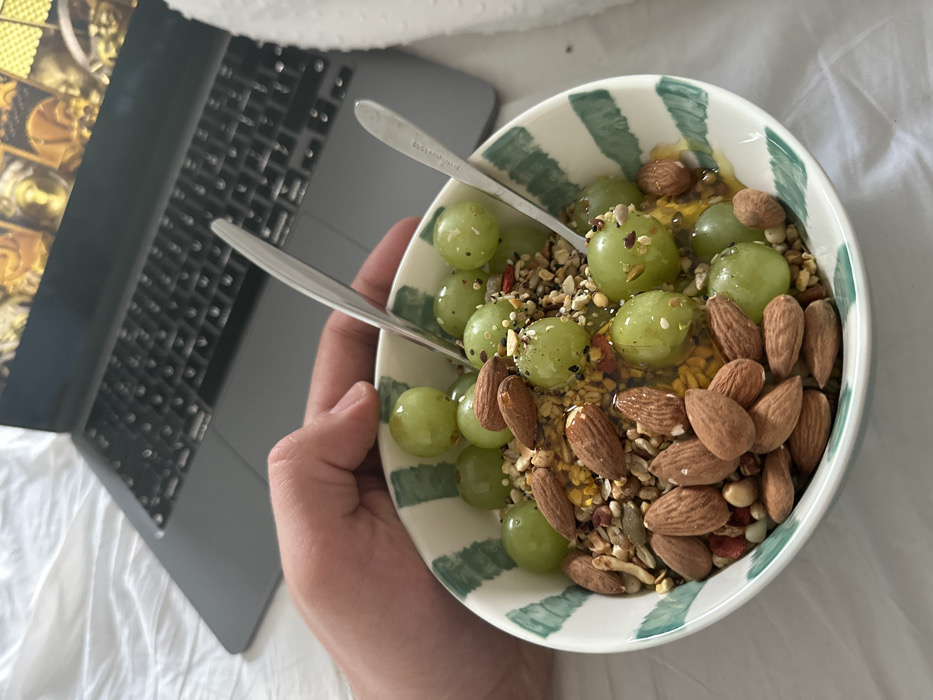 A close-up image depicts a person's left hand holding a white ceramic bowl with vertical dark green stripes. The bowl contains green grapes, whole almonds, a mixture of various seeds (including sunflower, sesame, and smaller black seeds), and a clear, golden liquid resembling honey or syrup. Two silver-colored spoons are resting within the contents of the bowl. In the background, out of focus, a dark grey laptop is positioned on white, wrinkled bedding. The laptop's screen displays a golden-yellow image with intricate patterns. The scene takes place indoors, on a bed, in the City of London, United Kingdom.