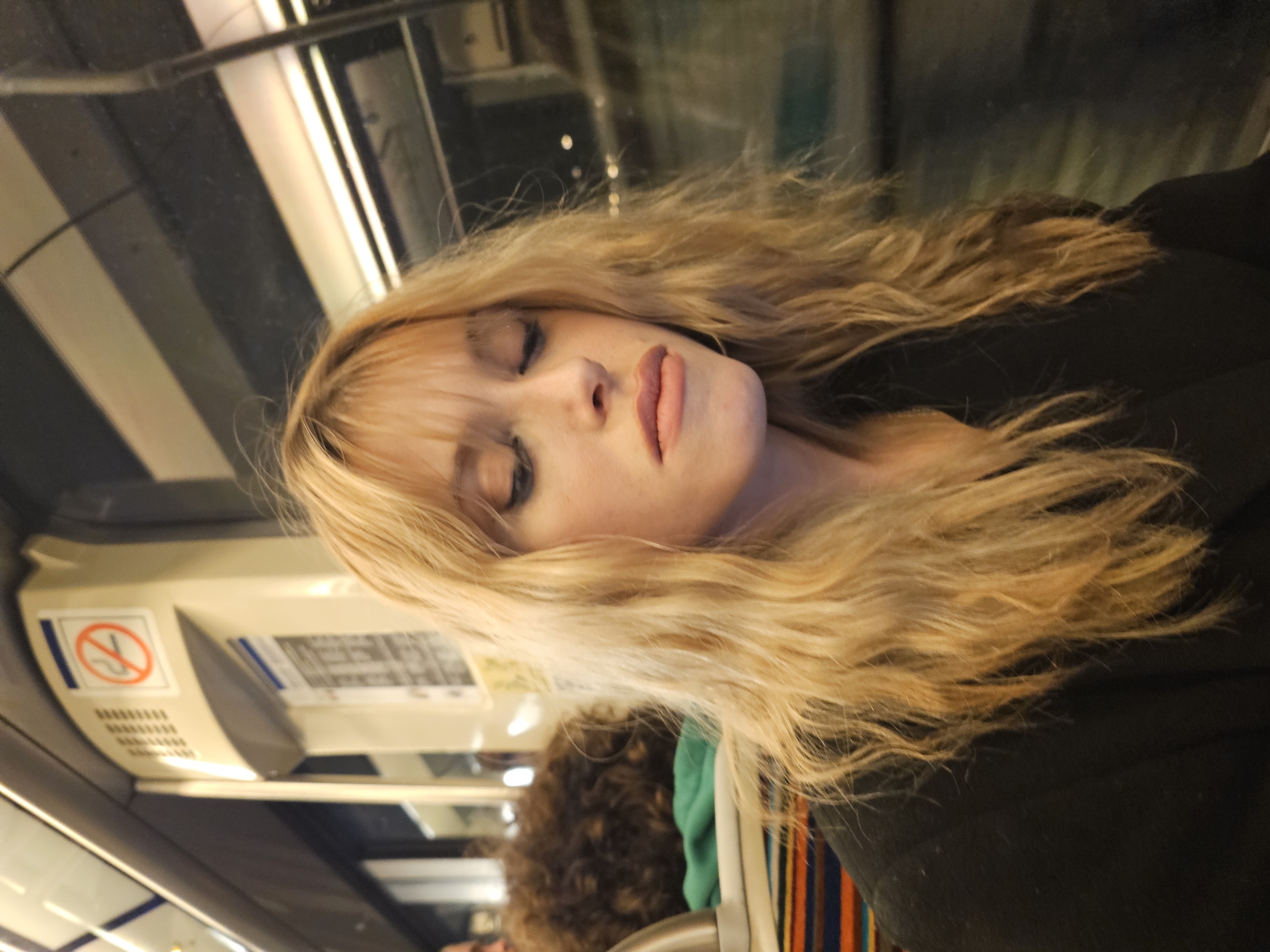 A woman is asleep on public transportation, possibly a train, in what appears to be the city of Paris, France. She has blonde hair, a fair complexion, and is wearing dark clothing. The background reveals elements typical of Parisian public transport such as signage and lighting fixtures. Additional people and objects are present, though out of focus.