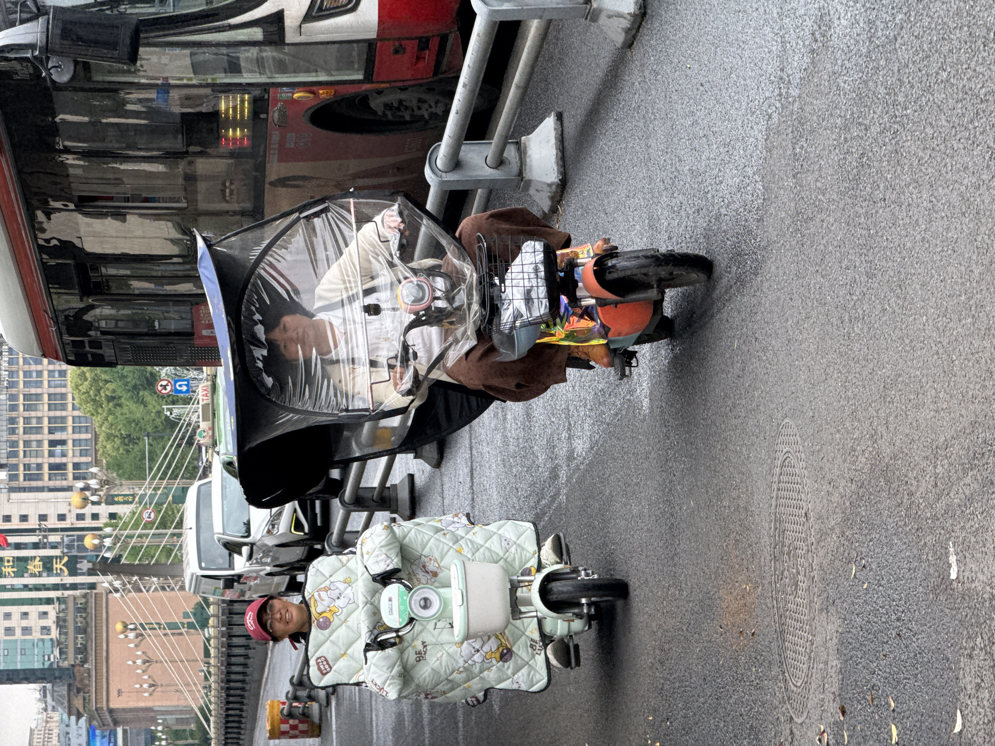 The image depicts two individuals riding electric bicycles on a wet road in Qingyang District, China. One person is wearing a red cap and is covered in a light green quilt with cartoon animal patterns while riding a small, light green electric bicycle. The other person is riding a larger electric bicycle with a protective black canopy, shielding them from the rain; they are wearing a white shirt and brown pants. A city bus is adjacent to them. Background includes city buildings, a bridge structure with ornate lamp posts, various vehicles, and street signs. The road is wet, reflecting the ambient light and suggesting recent or ongoing rain. A manhole cover is visible in the foreground.