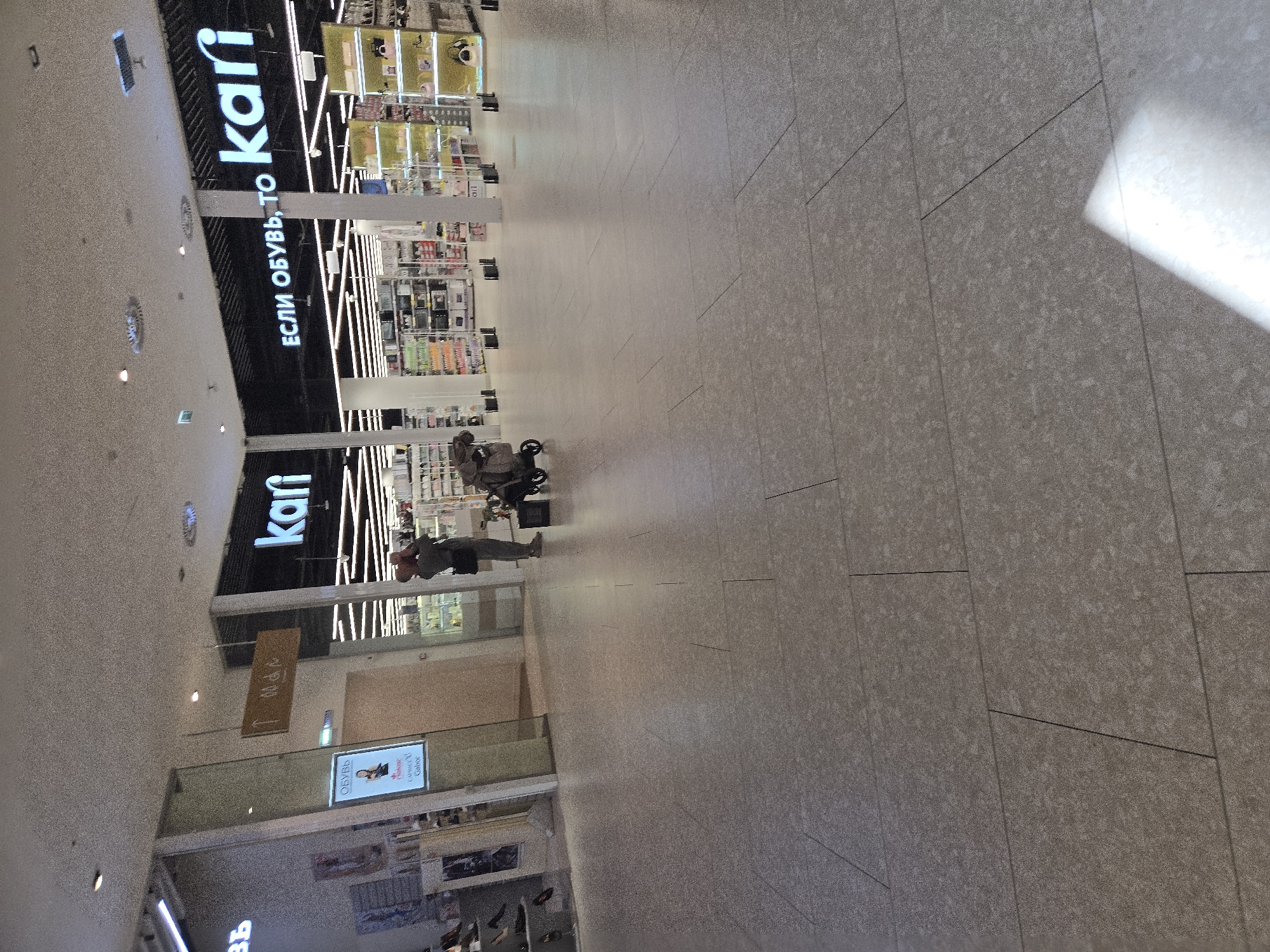 The image shows the interior of a retail store. A person is holding a baby while standing near a baby stroller. Shelves stocked with merchandise are visible in the background, suggesting a shopping environment. A sign above the shelves reads "KARI" in large letters, indicating the name of the store. Another sign in Russian says "ЕСЛИ ОБУВЬ, ТО" which translates to "If shoes, then". The floor is tiled with large grey tiles. The lighting is bright and appears to be artificial, coming from overhead fixtures. The overall impression is that of a busy retail space, likely a shoe store.