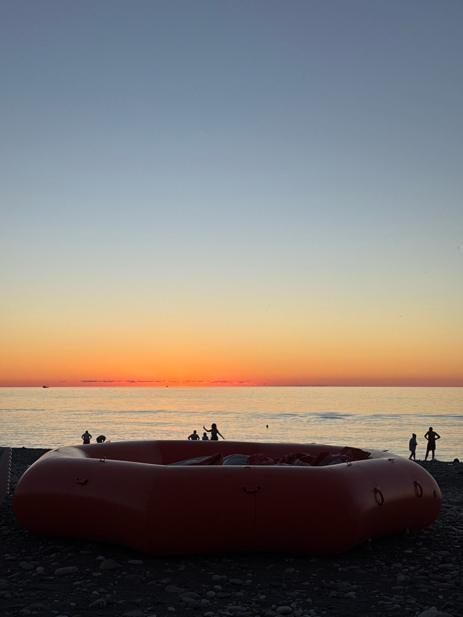 A red, inflatable raft rests on a pebble beach as a sunset casts an orange glow over the Black Sea. Silhouetted figures are visible on the beach and in the shallow water. The sky transitions from a pale blue to a vibrant orange near the horizon. The scene is set at Batumi, Georgia.