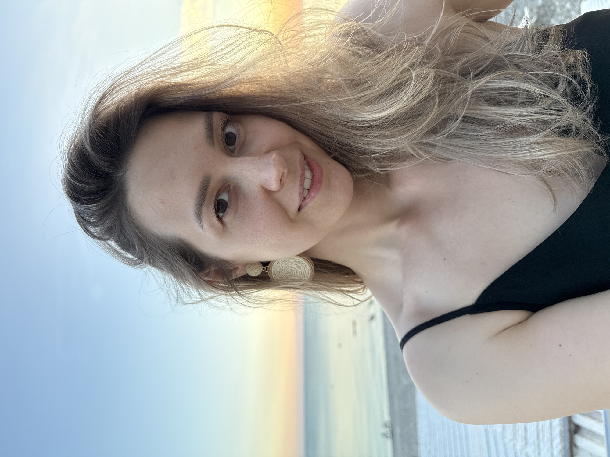 A light-skinned female individual is captured in a front-facing, waist-up portrait outdoors in Nebug, Russia, against a coastal sunset. She possesses long, windblown light brown hair with blonde highlights, parted on her left side. Her eyes are dark, and she exhibits a faint, closed-mouth smile directed towards the camera. She wears a black top with thin shoulder straps. A two-part earring, appearing to be made of woven light-colored material, is visible on her left ear, consisting of a circular stud and a larger oval dangle. The background features a calm body of water, likely the Black Sea, under a clear sky transitioning from deep blue at the top to lighter blue, yellow, and orange hues at the horizon, indicative of a sunset. The sun's light illuminates the right side of the subject's hair, creating a warm glow. Faint structures or a light-colored beach are discernible on the distant left shoreline.
