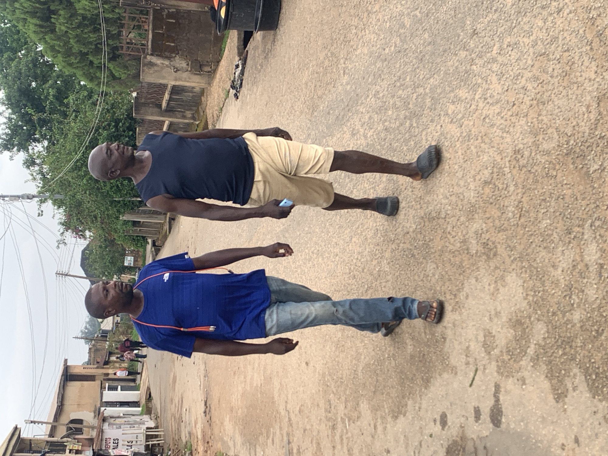Two adult black men are standing on a paved road in Jos, Nigeria. The man closer to the camera is wearing a blue shirt, jeans, and sandals, with headphones around his neck. The other man is wearing a dark blue sleeveless shirt, khaki shorts, and dark shoes, holding a blue smartphone in his left hand. The background shows buildings, trees, and utility poles, suggesting an urban residential or commercial area.