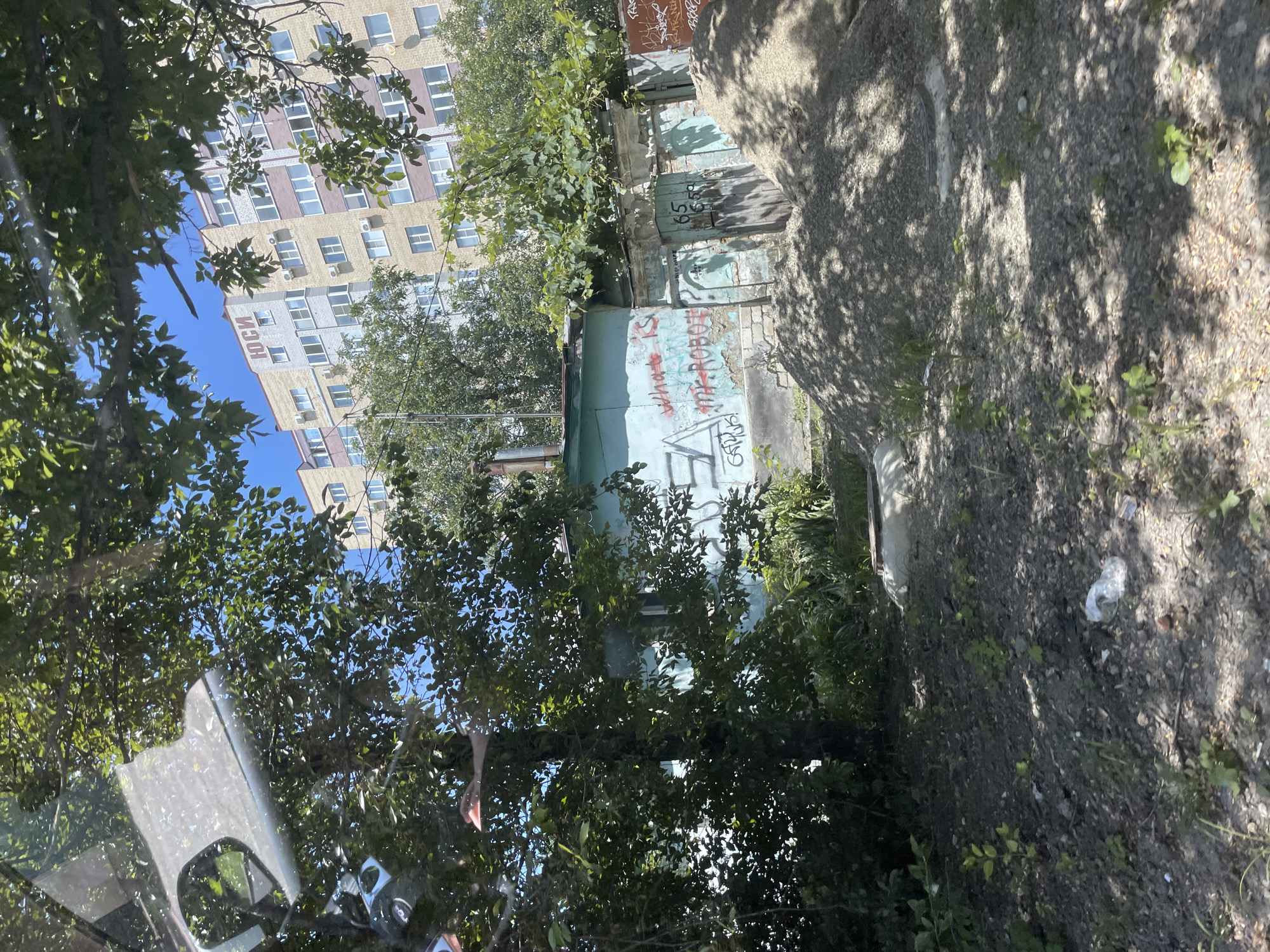 The image depicts a view from inside a vehicle, looking out at an urban scene. In the foreground, there is a pile of gravel, and beyond that, a low-lying, teal-colored structure with graffiti on its walls. The graffiti includes the words "what is" and "MR.ROBOT". A wooden door is visible on this structure, marked with the numbers "65".
Behind this structure, a multi-story apartment building with a light-yellow facade and brown trim is visible. Russian lettering, "ЮСИ" (YOUSI), is prominently displayed on the upper portion of this building. Trees with green foliage partially obscure the view of the buildings. The ground in the foreground is a mix of dirt, small stones, and sparse vegetation, with some visible litter, including a plastic bottle. The sky above is clear and blue. The scene appears to be an outdoor area in a residential part of Stavropol, Russia, possibly a courtyard or a construction site adjacent to residential buildings.